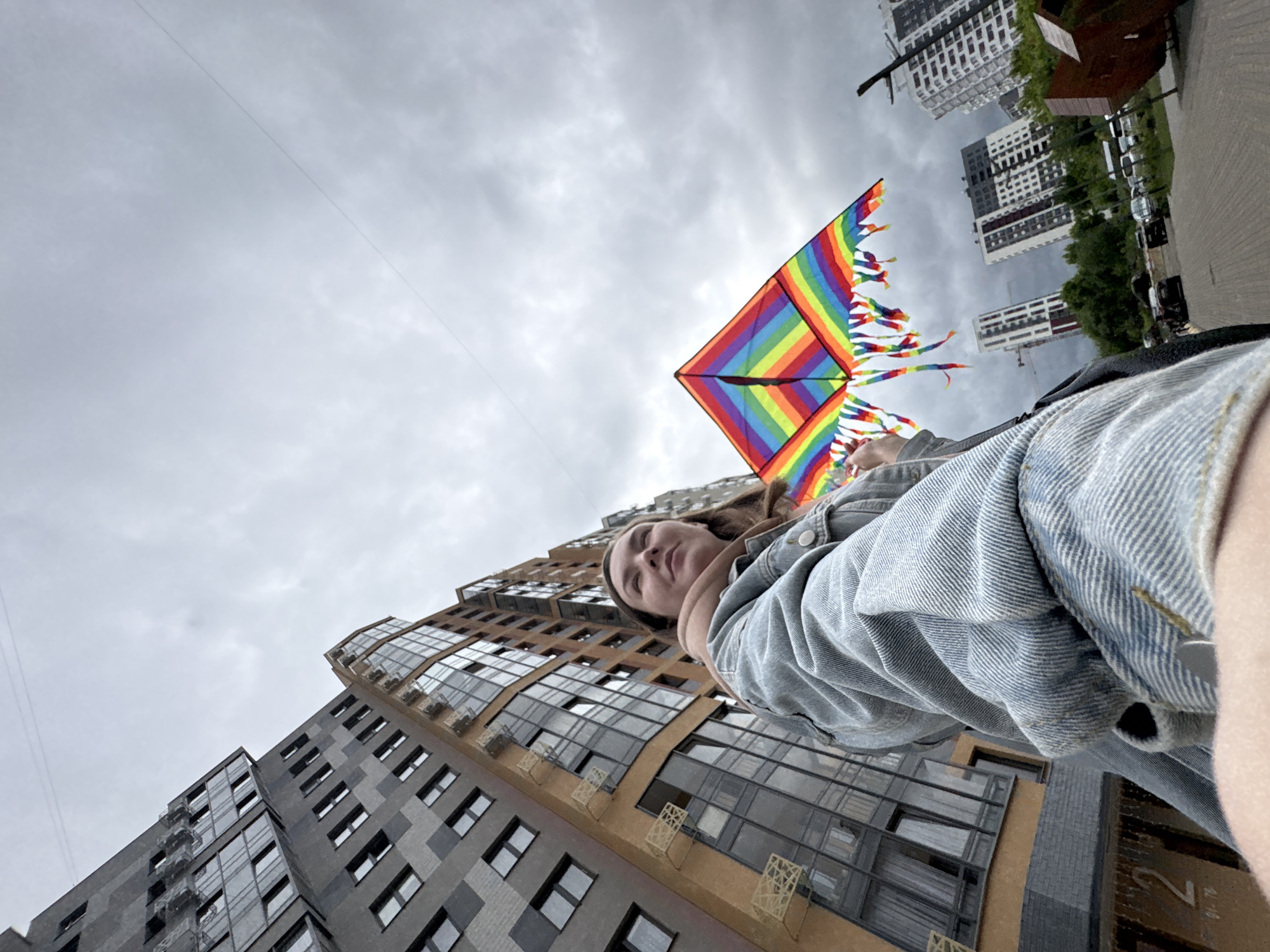 The image captures a young woman holding a rainbow-colored delta kite in an urban setting under an overcast sky. The woman is positioned in the lower-left, viewed from a low angle, and appears to be taking a selfie, with her right arm extended towards the camera. She wears a light blue denim jacket over a light brown hoodie, and her gaze is directed upwards and to her right. Her left hand is partially visible, holding the string of the kite. The kite, positioned in the mid-right, features distinct horizontal stripes of red, orange, yellow, green, blue, indigo, and violet, with a multi-colored fringe along its bottom edge.
The scene is situated within the Omega Residential Complex (ЖК Омега) in Izhevsk, Russia. Two modern multi-story residential buildings dominate the left side of the frame. The building in the immediate foreground exhibits a facade with dark gray panels accented by lighter gray geometric patterns and numerous rectangular windows. Its lower section features a brown facade with large glass windows, and specific numerical markings "2" and "91-79" are visible on its lower exterior wall. Adjacent to it, another tall residential building is visible with an orange-brown facade and extensive glass balconies and windows. In the background to the right, several other modern high-rise buildings are discernible amidst a line of green trees. A paved ground area, likely a road or pedestrian path, is visible in the lower right, with parked vehicles and a dark, angular object resembling a large waste container. The sky is uniformly gray and heavily clouded, indicating an overcast day.