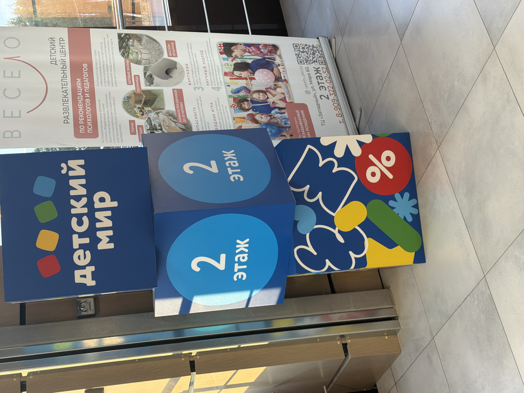 The image shows a retail environment, likely a shopping mall, with a brightly colored display for a children's entertainment center. The display features prominent signage with the name "ДЕТСКИЙ МИР" (Children's World) and various promotional materials. A large blue sign with "2 этаж" (2nd floor) in white text is a focal point. A banner behind the display provides information about the entertainment center, including its services and a picture of children at a party. The floor is tiled with light gray tiles. The scene is well-lit, with sunlight casting shadows on the display. There are no people visible in the immediate foreground. The environment appears clean and organized, typical of a commercial establishment. The city and specific location are unknown, but the context suggests a public space within a shopping complex.