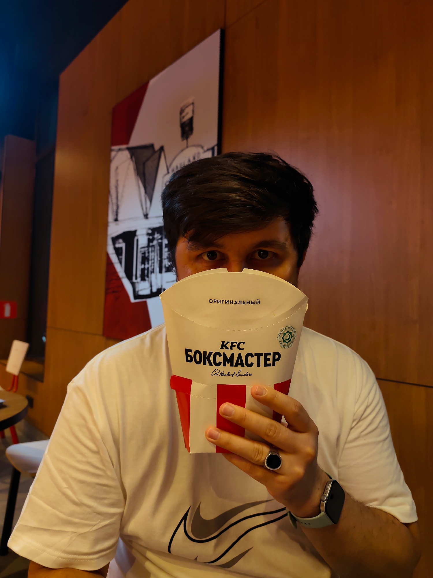 A man in Fergana City, Uzbekistan, is holding a KFC box in front of his face. He is wearing a white t-shirt and has a ring on his left hand and a smartwatch on his right wrist. The background consists of a wooden wall and a framed picture.