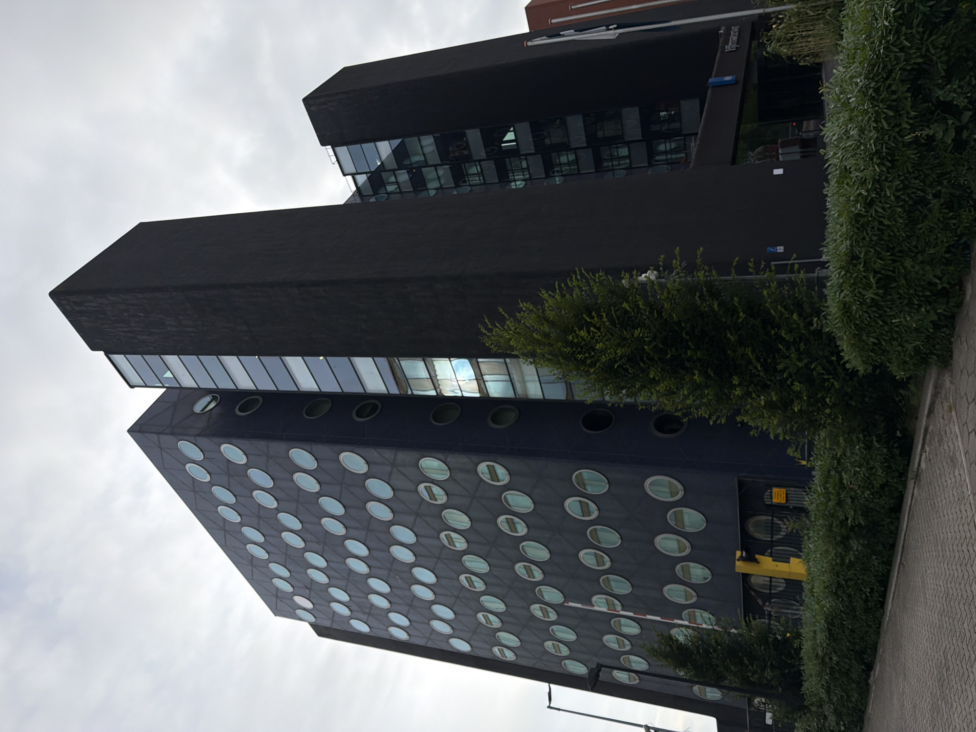 The image shows a large, dark-colored modern building with a distinctive facade featuring rows of circular windows. The building appears to be an office building or hotel, with a contemporary architectural style. The facade on the left side of the building is dark blue and has a grid pattern with many round, light-blue tinted windows. The right side of the building is a stark, black, monolithic structure that contrasts with the other side. Between these two sections, there is a glass facade that reflects the sky and surrounding environment.
In front of the building, there is a well-maintained green hedge. A paved area, likely a sidewalk or parking lot, is visible in the foreground. A street lamp and a retractable barrier are present near the entrance of the building. The sky is overcast and cloudy, indicating a possibly temperate climate.
This building is the former headquarters of the Dutch Tax Administration (Belastingdienst) and is located in Rijswijk, Netherlands. The architectural design is quite unique and easily recognizable.