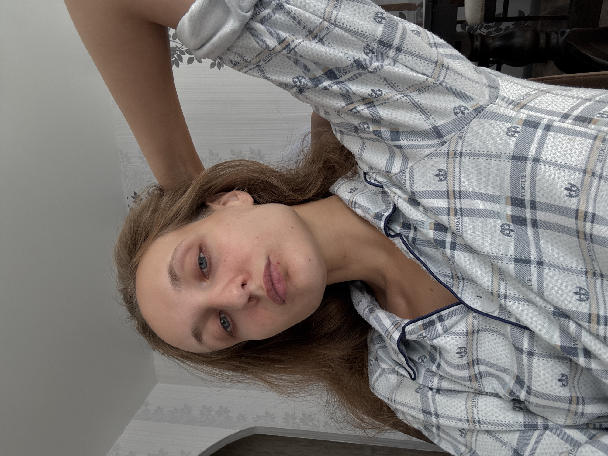 A young woman with long, light brown hair is pictured from the chest up, indoors, with her right arm bent behind her head. She is wearing a plaid pajama top with navy blue trim and a button-up front. Her eyes are blue and she is looking directly at the camera with a neutral expression. The background consists of a wall with floral wallpaper and a portion of an arched doorway to her left. To the right, part of a dark wooden chair and table are visible. The lighting appears to be natural, coming from a window or overhead source. The image does not provide any specific information about the location within Sharkan, Russia.