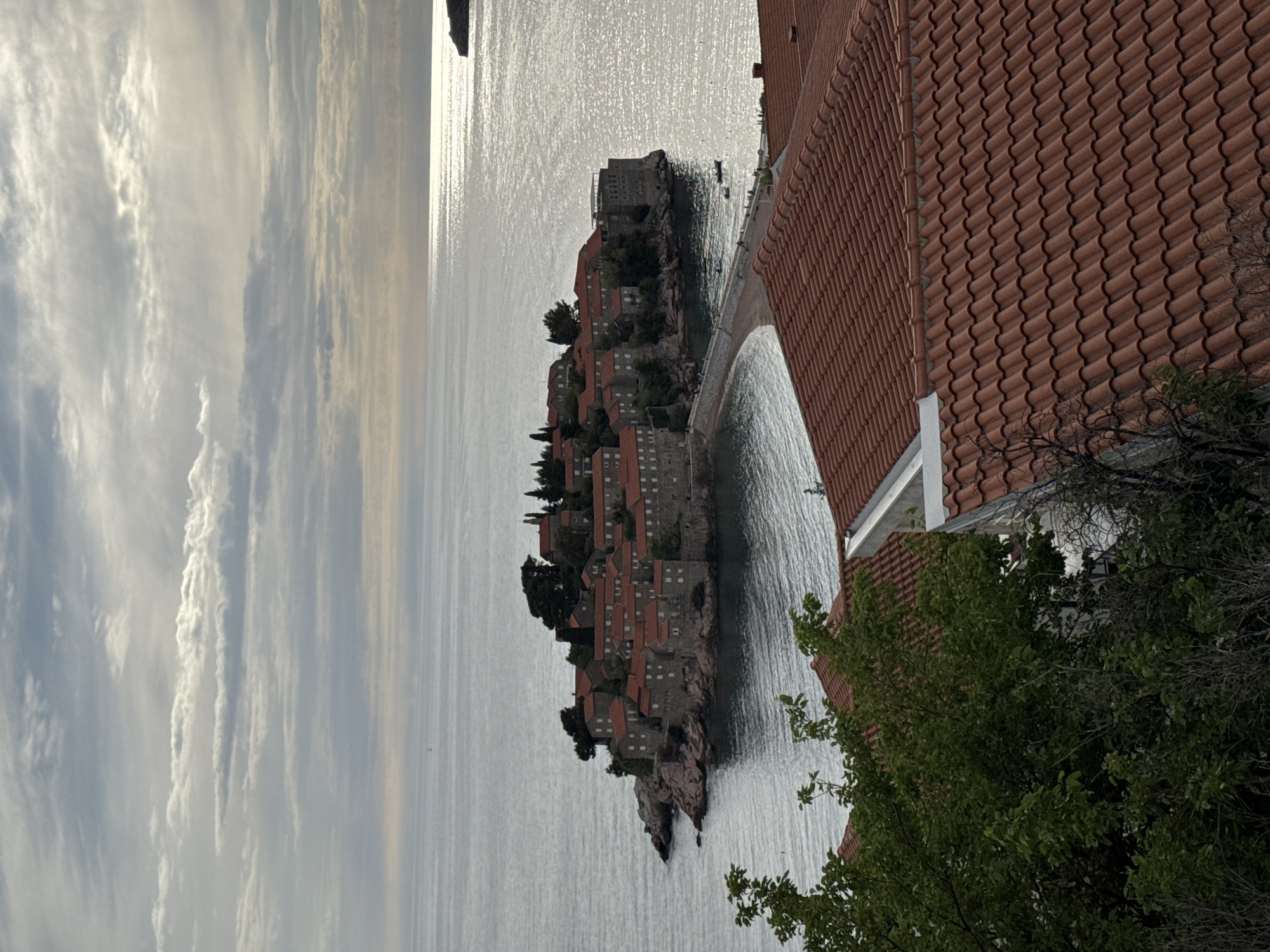 The image depicts a view of the island of Sveti Stefan in Montenegro. The island, covered in buildings with red roofs, is connected to the mainland by a narrow isthmus. The Adriatic Sea surrounds the island, reflecting the sunlight. To the right of the island, there are buildings with terracotta tiled roofs and green trees. The sky is filled with streaks of clouds. The photograph appears to be taken from an elevated perspective.