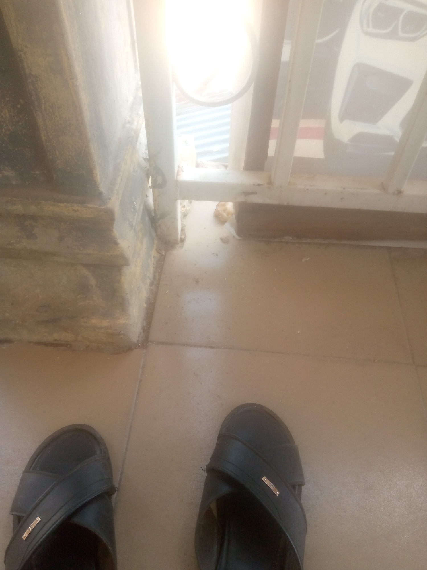 The scene depicts a ground-level view in an outdoor or semi-enclosed area in Jalingo, Nigeria. In the immediate foreground, the feet of an individual wearing black slip-on sandals with small gold-colored accents are visible on a light brown tiled floor. The tiles appear dusty. To the left, a multi-tiered concrete pillar, exhibiting signs of age and discoloration, rises vertically. A white metal railing, composed of vertical and horizontal bars, extends from the right side of the frame towards the center, separating the foreground from the background. Accumulations of dirt and small fragments of debris are present on the tiled floor at the base of both the concrete pillar and the metal railing. Beyond the railing, the background is heavily overexposed by a bright light source, likely direct sunlight. Through this glare, the front portion of a white vehicle, possibly a car, is partially visible, either as a reflection or a large printed image. Faint corrugated metal structures are also discernible in the heavily illuminated background.