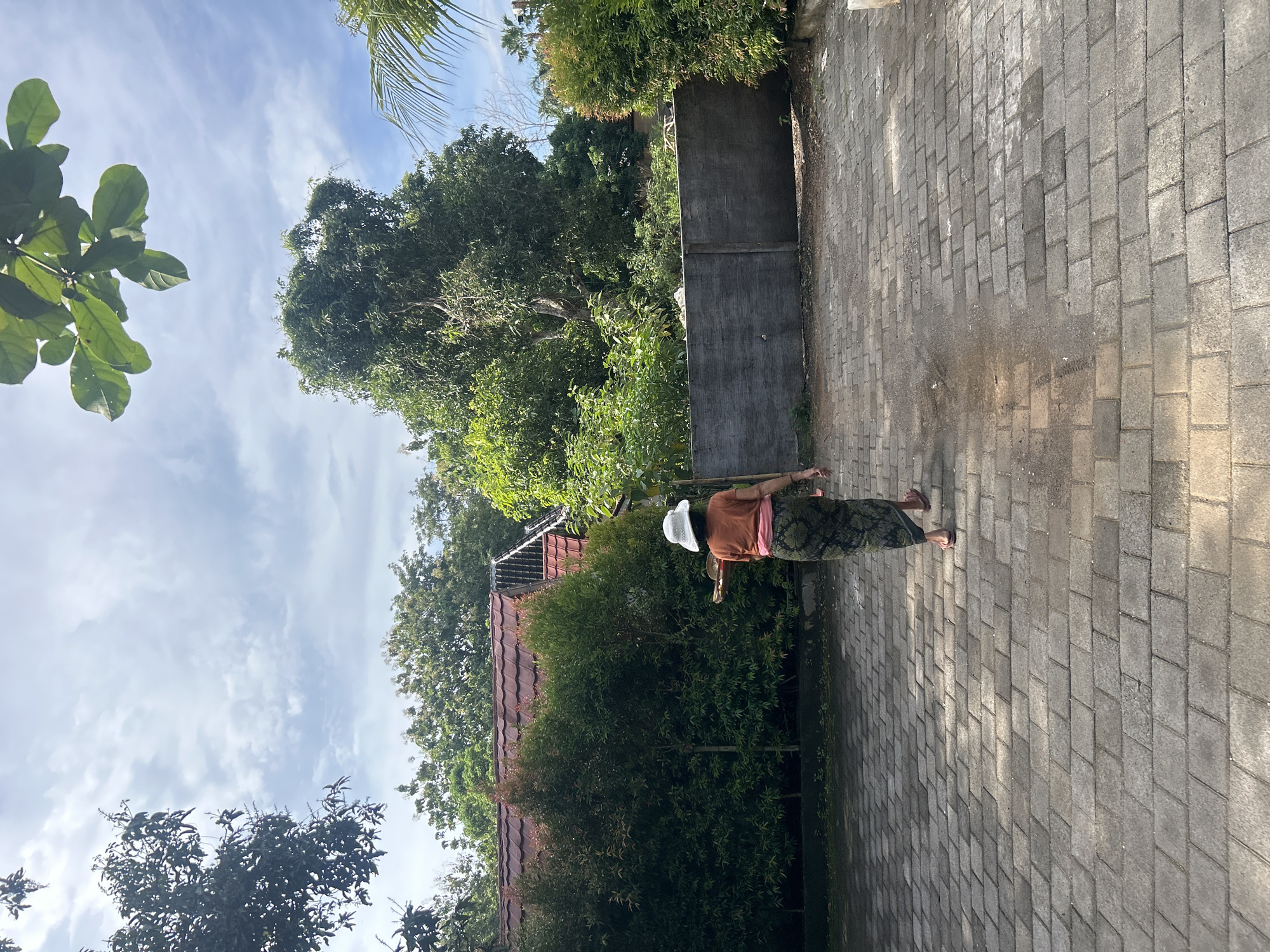 A person wearing a brown shirt, patterned skirt, and a white hat stands on a brick-paved ground. The sky is blue with scattered clouds. Trees and foliage surround the area, with a wall and a red-tiled roof visible in the background. The image seems to be taken in Kuta Selatan, Indonesia, based on the environment.