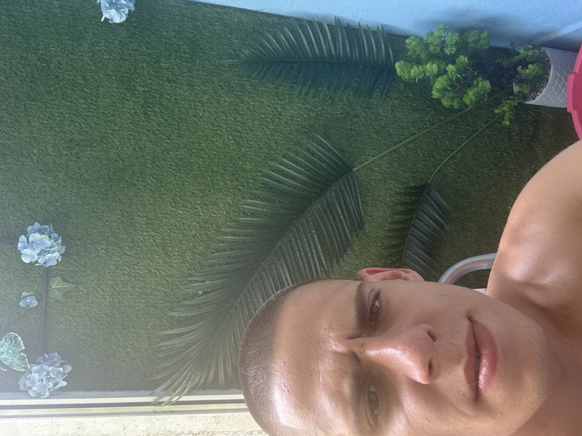 A close-up, selfie-style photo shows a young man with a buzz cut and a furrowed brow, looking directly at the camera. He is shirtless, and the background consists of a green, textured wall, possibly artificial grass, adorned with artificial blue flowers and palm fronds. A small potted plant with green foliage sits in the lower right corner. The lighting suggests it is daytime, possibly outdoors or near a window, with some harsh shadows. The man's expression implies concern or displeasure. The scene does not provide any specific indicators of its location within the city of Sofia, Bulgaria.