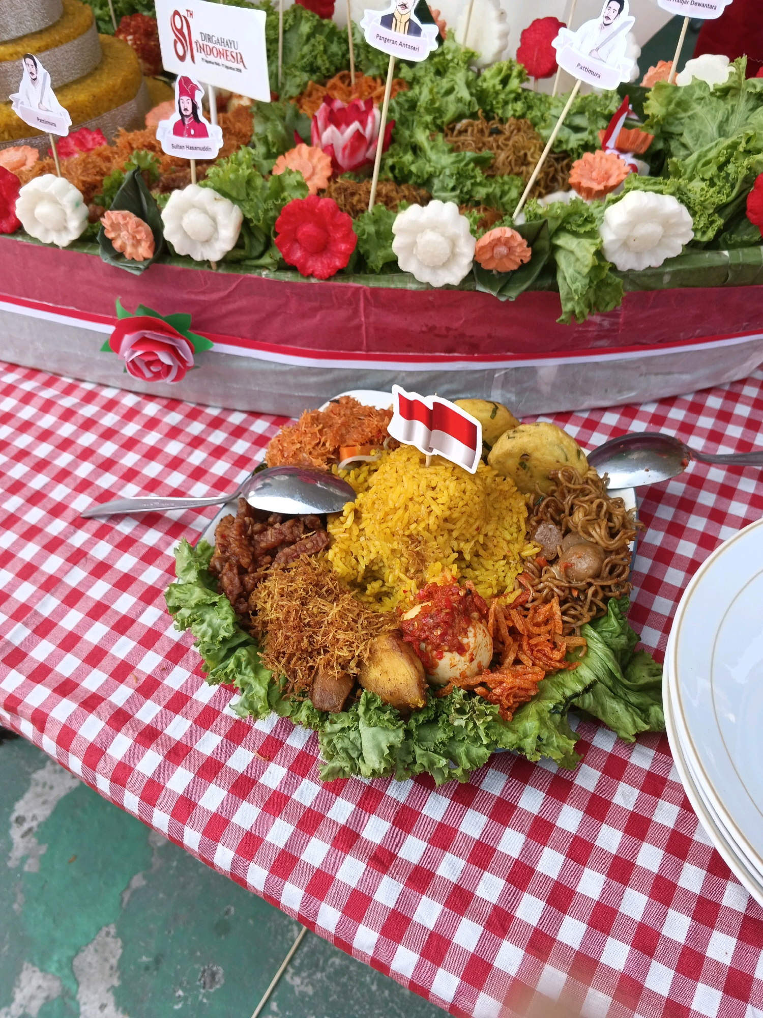 A high-angle, medium shot captures a close-up of a platter of Indonesian food arranged on a red and white checkered tablecloth, likely for a celebration in Margahayu, Indonesia. The platter, which is white, is filled with a colorful assortment of dishes. In the center, bright yellow rice is piled high and topped with a miniature Indonesian flag. Surrounding the rice are various side dishes including fried noodles with meatballs, shredded chicken, tempeh, and hard-boiled eggs coated in a spicy red chili sauce. Lettuce leaves form a bed for the food, and decorative carved carrots and white floral-shaped food items are scattered throughout. In the background, a tiered cake or traditional tumpeng is visible, adorned with small flags featuring images of historical figures. The overall scene suggests a festive and patriotic occasion.