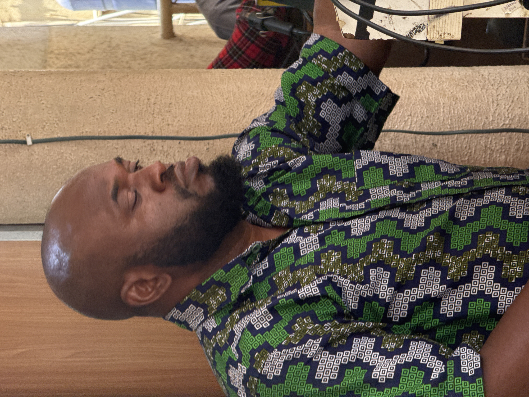 An image, rotated 90 degrees clockwise, shows a man with dark skin, a shaved head, and a neatly trimmed dark beard, apparently resting or in contemplation. His eyes are closed, and his expression is serene. He is wearing a short-sleeved shirt with a distinctive geometric pattern of squares in shades of green, dark blue, white, and olive/khaki.
The setting appears to be an indoor environment, possibly a workshop or storage area, within Abuja, Nigeria. In the background, there are large, light brown, textured rolls that resemble carpets or fabric, positioned vertically. A dark cable stretches horizontally across these rolls. Above the man's head, parts of a metal structure and some other materials, including a piece of red plaid fabric, are visible. In the upper right corner, attached to the metal structure, is a piece of paper or cardboard with handwritten text that includes what appears to be "AD" or "A.D.", possibly "ECA" or "ECG", and the number "664". The lighting suggests it is daytime, with potentially additional artificial illumination. No specific activity beyond the man's resting posture is depicted.