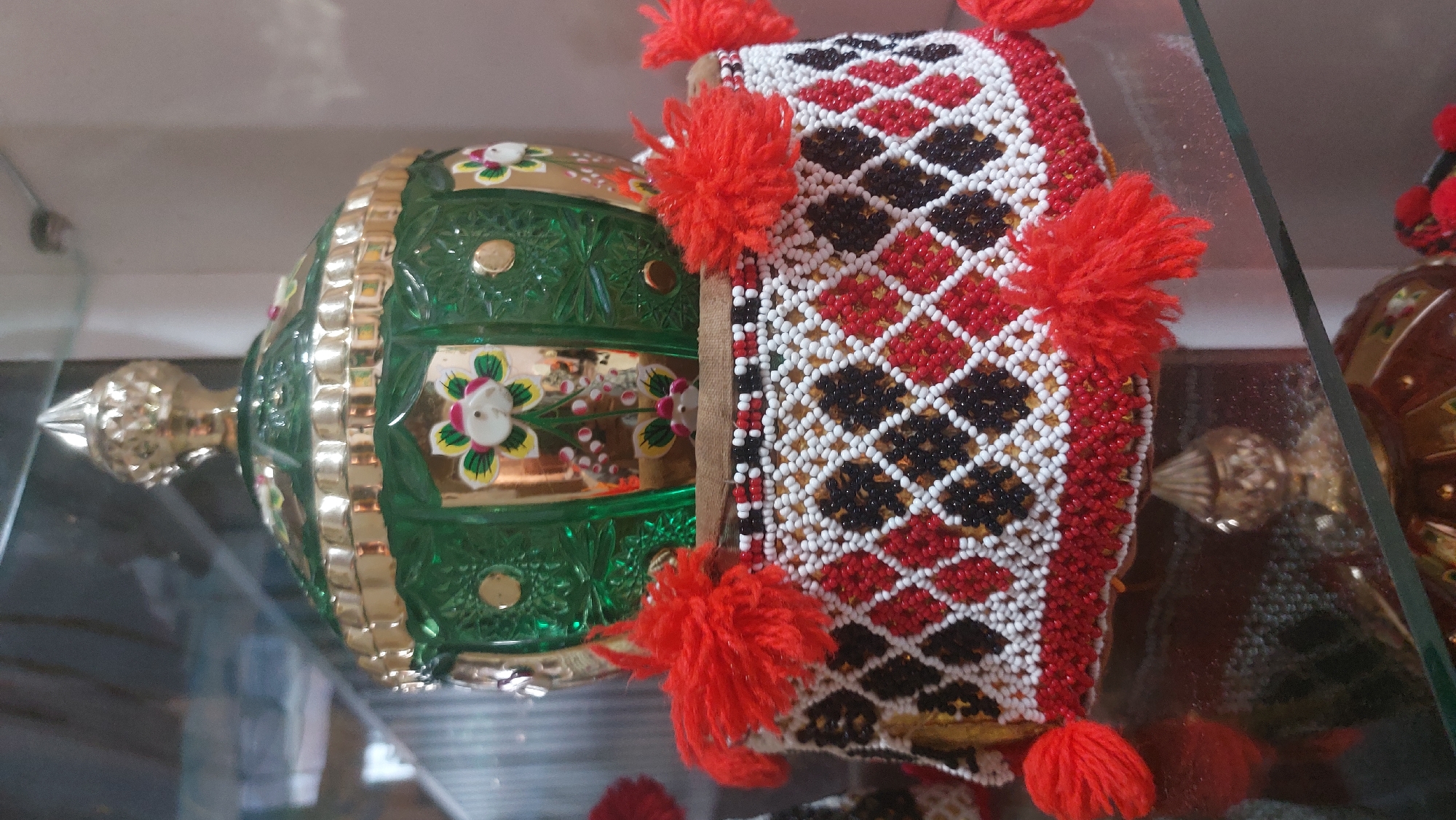 A close-up shot showcases a decorative, beaded cylindrical object with red, black, and white diamond patterns. Red pom-poms are attached around its top edge, and it supports a green, faceted glass dome adorned with gold trim and floral motifs. The object appears to be displayed on a reflective surface, possibly a glass shelf, within a dimly lit environment. The background is blurred, with hints of shelving and possibly other decorative items. This object is likely part of a collection of cultural artifacts exhibited in a museum or gallery in the City of Medan, Indonesia, perhaps at the Museum of North Sumatra.