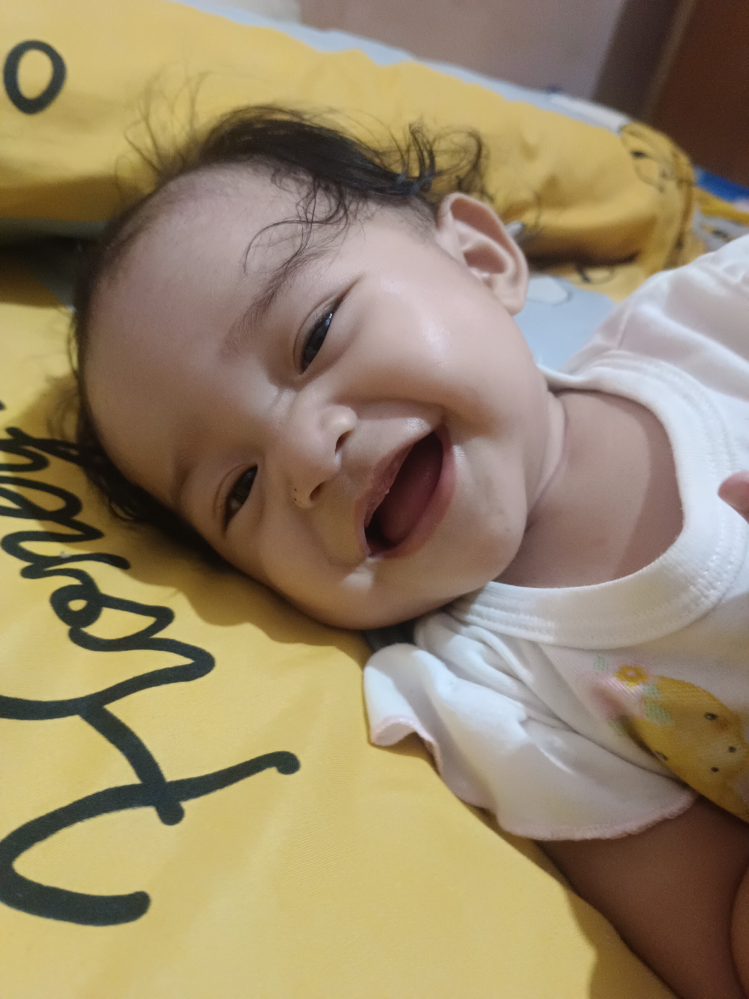 A close-up shot captures a smiling baby lying on a yellow surface with black lettering. The baby is wearing a white onesie with a floral pattern. The baby's head is tilted to the left, and their eyes are open and looking towards the camera. The baby's mouth is open, revealing a wide smile. The background is blurred, but appears to be a bed with yellow bedding. The lighting is soft and warm, suggesting an indoor setting. The image was taken in Kawua, Indonesia.