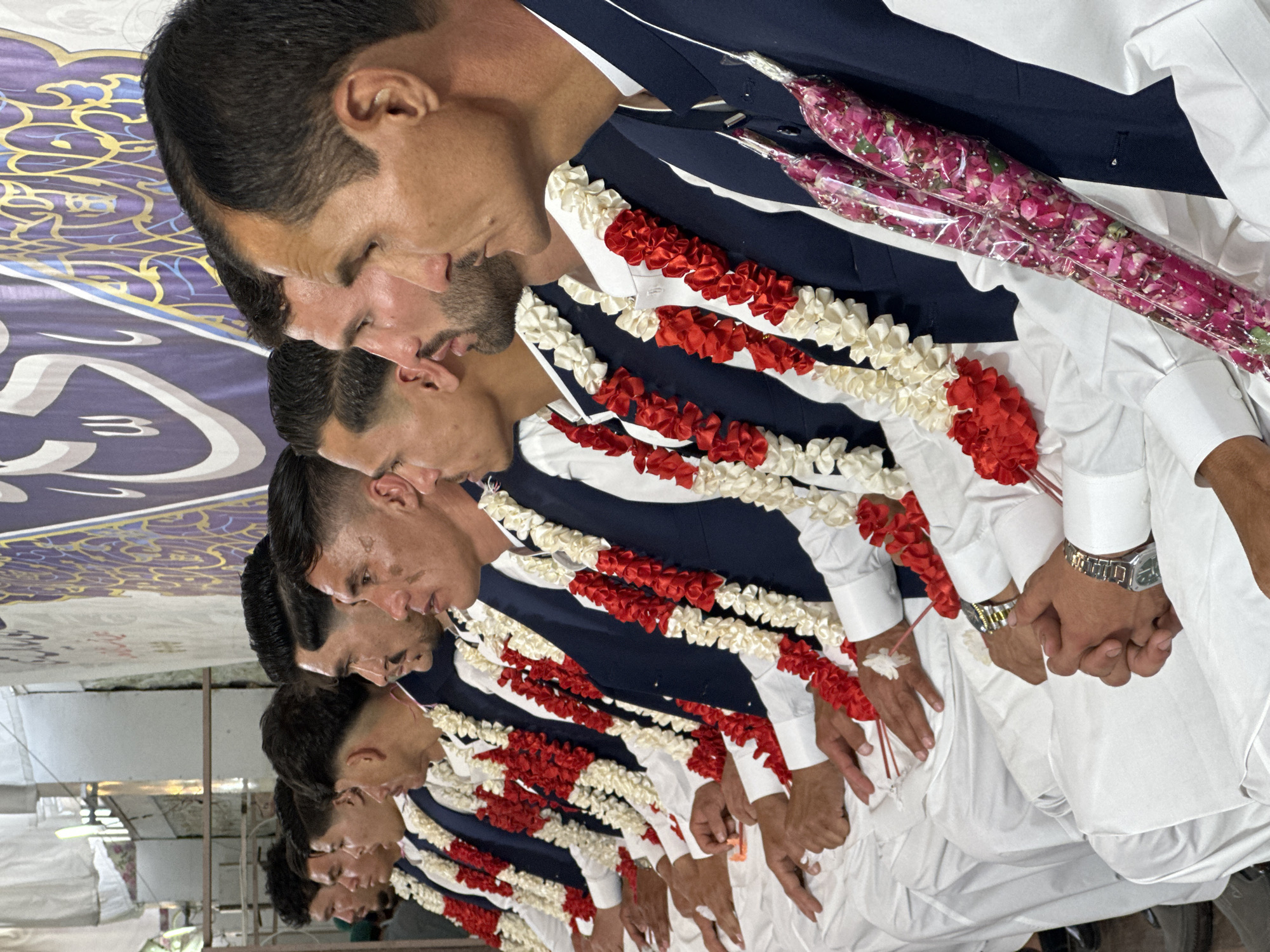 A row of at least seven men dressed in traditional Pakistani attire stand in a line, facing the same direction. Each man wears a white shalwar kameez, a dark blue waistcoat, and a garland of red and white flowers around his neck. The men are holding hands. In the background, there is a decorated fabric or wall hanging with gold and blue patterns. The scene appears to be in Quetta City Tehsil, Pakistan.