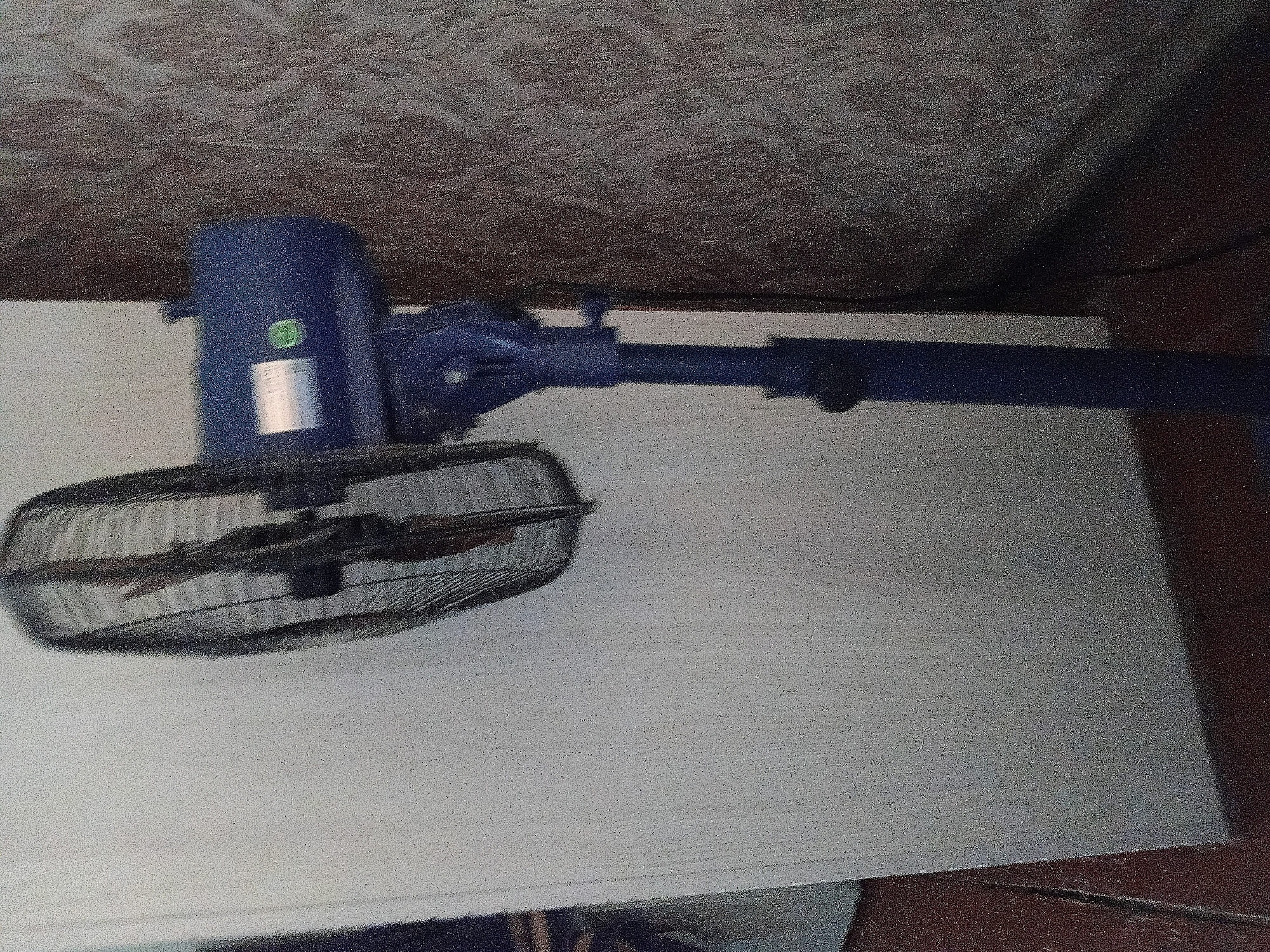 The image shows a blue oscillating fan standing on a dark floor next to a textured, patterned wall. The fan is positioned in front of a plain, light-colored surface, possibly a wall or screen, which occupies the left side of the frame. The fan has a protective grill covering its blades and a central motor housing with a label and a green circular sticker. The stem of the fan is also blue, and it sits on a circular base. The lighting in the scene appears to be dim, casting shadows. There are no people visible in the image. This scene does not provide any information to determine its location within the city of Kamen-na-Obi, Russia.