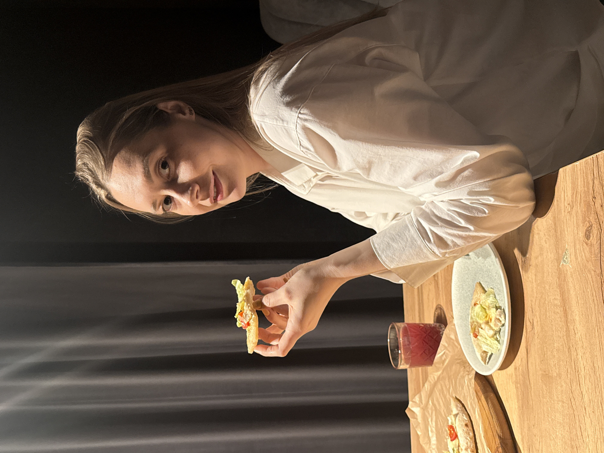The image shows a person with fair skin and long blonde hair, wearing a white collared shirt, seated at a light brown wooden table. The person is holding a piece of food resembling a taco or flatbread with lettuce and possibly other fillings. On the table, there's a white plate with a similar dish, a glass of red liquid, and what appears to be more of the flatbread food items on a piece of parchment paper. The background is dark, with draped grey fabric. The location is not identifiable from the image.