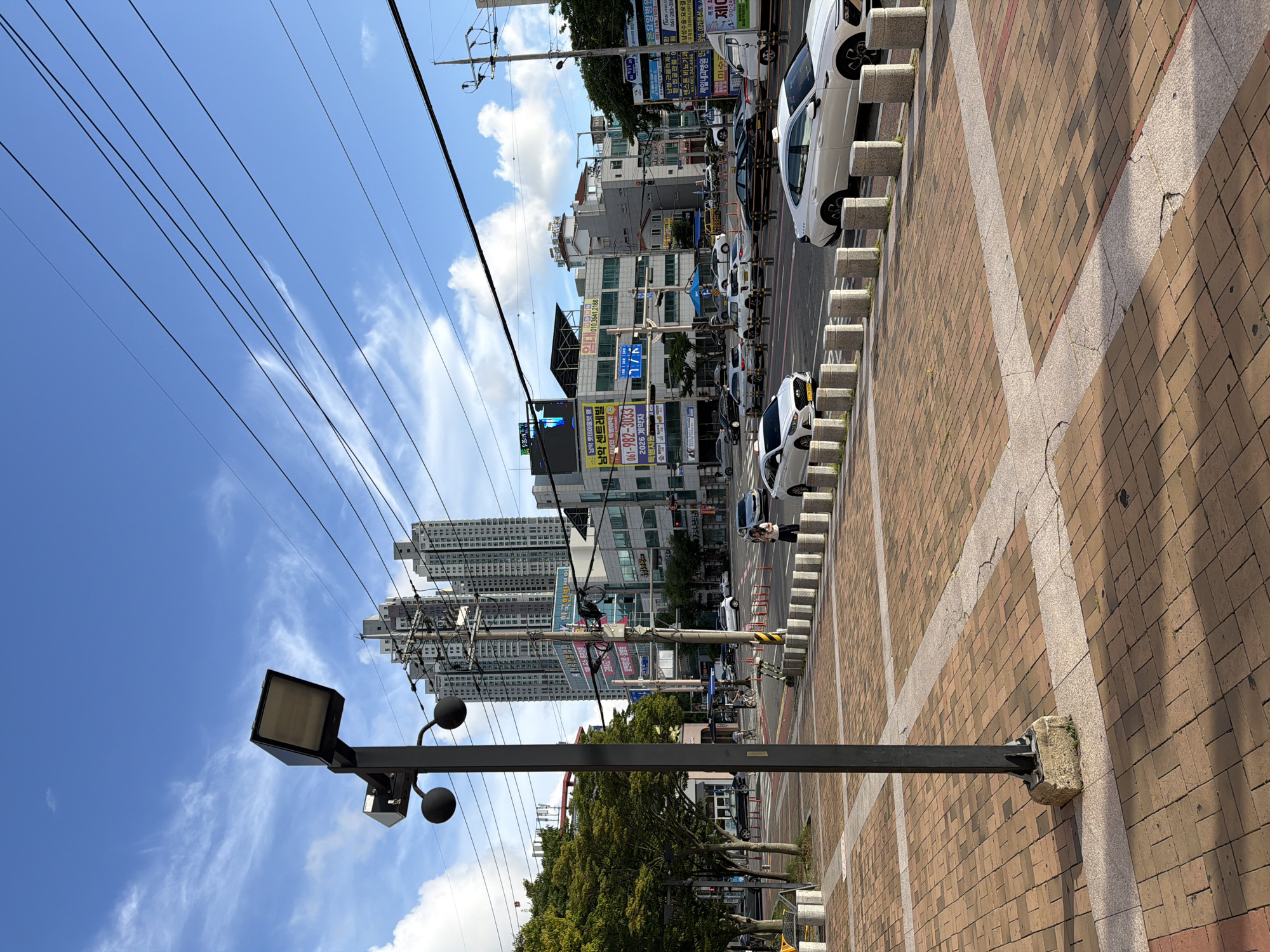 The image depicts a street scene in Mokpo-si, South Korea, on a clear day with a blue sky and scattered clouds. A lamppost with two spherical lights and a rectangular fixture stands in the foreground, casting shadows on the brick-patterned sidewalk. In the midground, a street with multiple lanes of traffic is lined with concrete bollards. Several white cars, including two taxis, are visible on the road. Buildings of various sizes and architectural styles line the street, some featuring signage with Korean text. In the background, tall apartment buildings rise against the sky. The environment suggests a commercial or residential urban area.