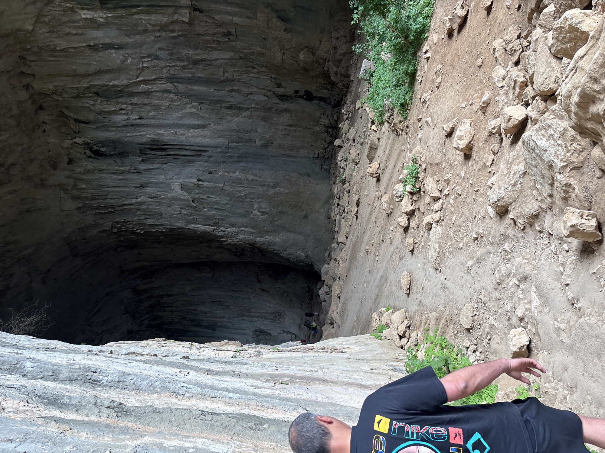 The image shows the entrance to the Devetashka Cave near Karlukovo, Bulgaria. A person wearing a black Nike t-shirt lies prone on the cave's edge, with their head and arm visible in the foreground. The vast, dark opening of the cave dominates the left side of the frame, revealing layered rock formations within. To the right of the cave entrance, a steep, rocky slope is visible, covered with loose soil, stones, and sporadic vegetation. The cave opening is massive and appears to descend significantly into the earth.