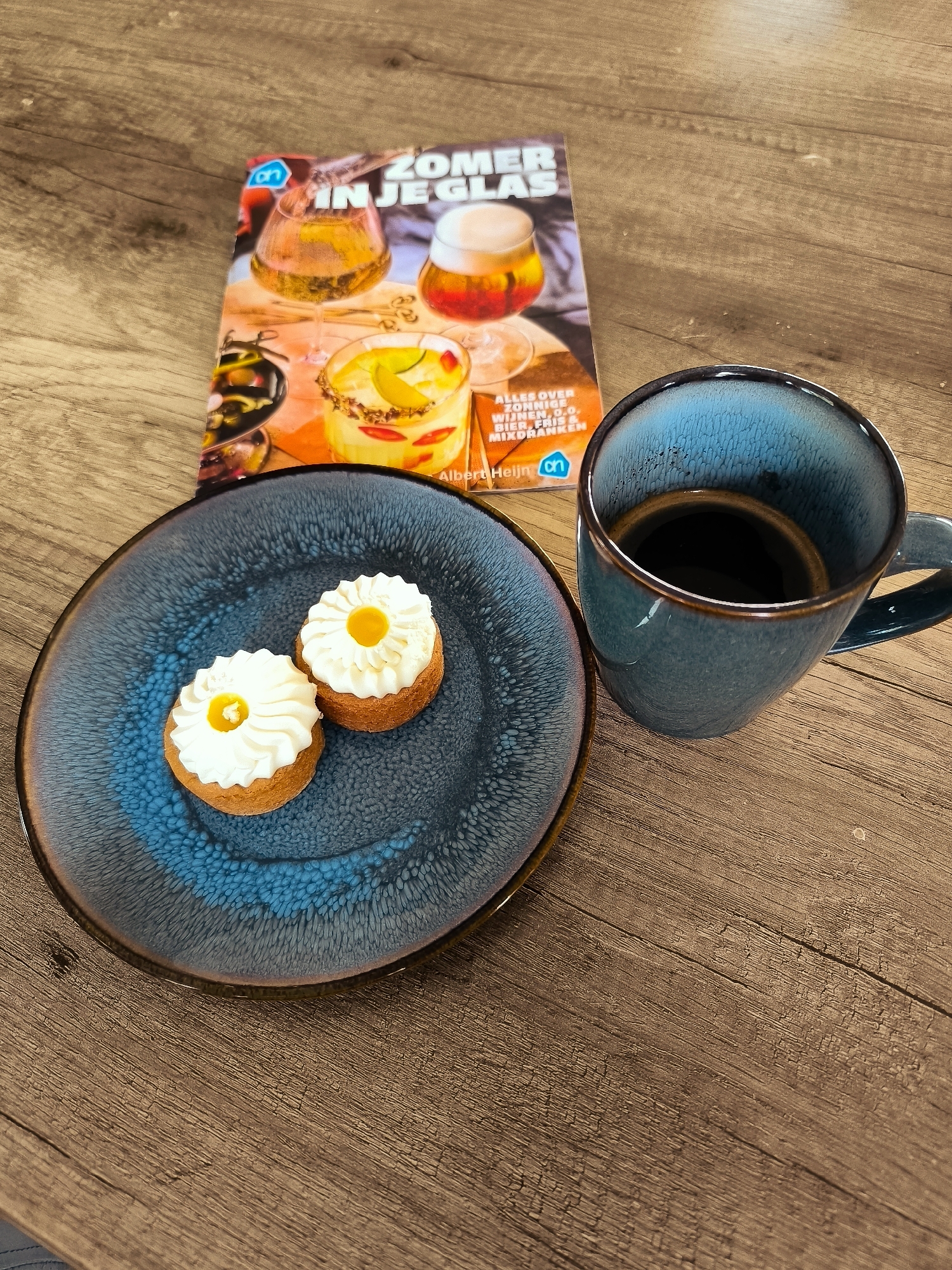 A close-up, high-angle, outdoor shot on a wooden table shows a book titled "ZOMER IN JE GLAS" (Summer in your glass), a blue speckled plate with two small pastries topped with white frosting and yellow centers, and a blue ceramic mug filled with dark coffee. The book is positioned to the left of the coffee mug and plate. The pastries are arranged side-by-side on the plate. The coffee mug is to the right of the plate. The wooden table has a visible grain pattern. The scene appears to be in Almere, Netherlands, likely on a balcony or patio given the lighting and ambiance suggested by the "summer" theme of the book.