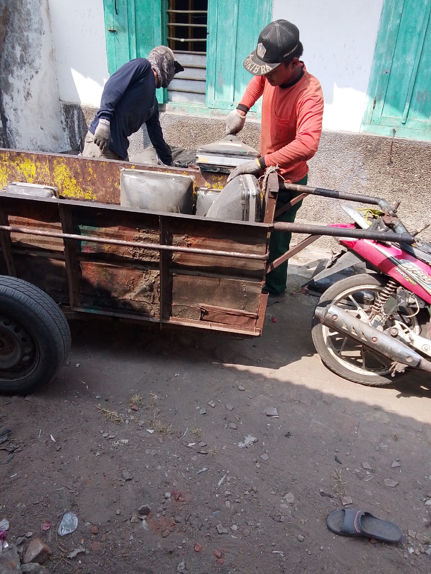 Two individuals, likely sanitation workers, are engaged in loading debris into a dilapidated, rust-colored trailer attached to a red motorcycle. The individual on the left, wearing a dark long-sleeved shirt, light pants, and a patterned head covering, is bent over the trailer, appearing to place items inside. The individual on the right, sporting an orange long-sleeved shirt, green pants, and a black baseball cap, is also working with the trailer's contents, holding what appears to be a gray object. Both individuals are wearing work gloves. The trailer, showing significant signs of wear and tear with peeling yellow paint and exposed metal, is positioned on a dirt ground. To the right of the trailer, the rear of a red motorcycle is visible, with its wheel and exhaust pipe in the frame. The background features a white wall with a green-shuttered window, suggesting an outdoor setting, possibly in Bulurejo, Indonesia, given the context provided. The ground is a mix of dirt, small stones, and scattered debris, including a single black sandal visible in the lower right portion of the image. The scene is illuminated by bright sunlight, casting shadows.