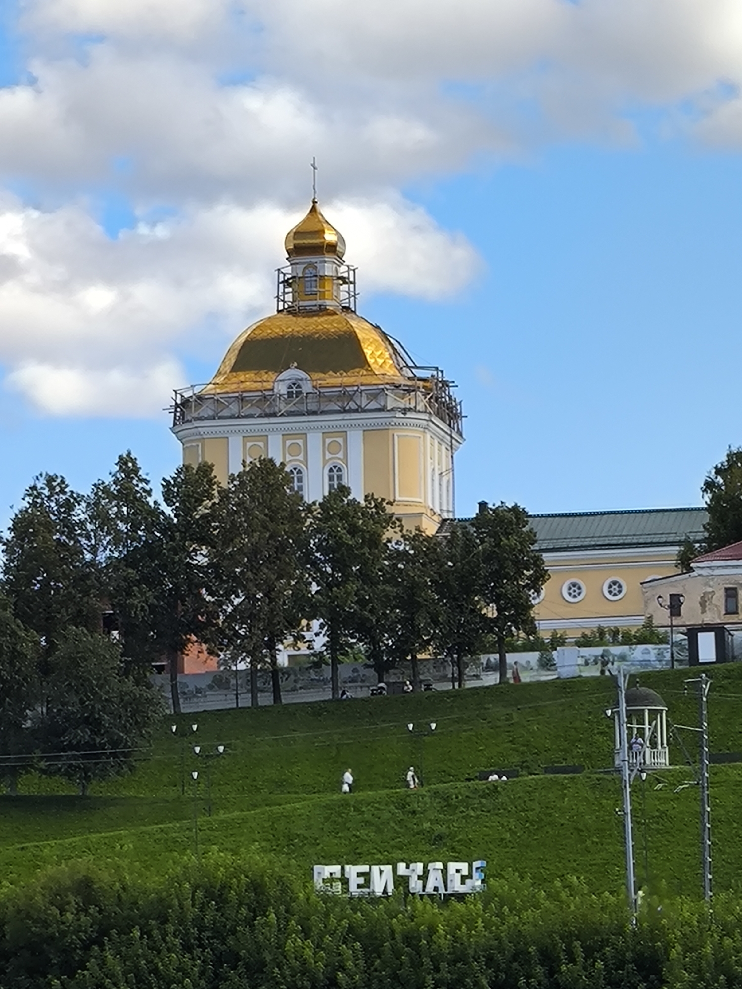 The image depicts a bright, overcast day in Perm, Russia, with a clear blue sky peeking through scattered white clouds. In the background, a grand, yellow building with a golden dome, likely a church or a significant historical structure, is prominently displayed. Scaffolding is visible around the dome, suggesting ongoing renovation or construction. A row of lush green trees lines the foreground, obscuring the lower portion of the building and creating a natural frame.
In the middle ground, a grassy slope descends towards the viewer. Several figures, appearing as small dots from this distance, are scattered across the slope. Some are walking, while others seem to be standing or interacting. To the right, a small, white gazebo with a dark roof is situated on the slope, with at least one person visible inside. A tall, slender metal structure, possibly a utility pole or a lamppost, stands to the right of the gazebo, extending upwards.
At the bottom of the image, large white block letters spelling out "ПЕРМЬ" (Perm) are visible, set against a backdrop of dense green foliage. The lettering is a prominent landmark, likely indicating the city's name. The overall scene conveys a sense of civic pride and historical significance, set against a backdrop of natural beauty.