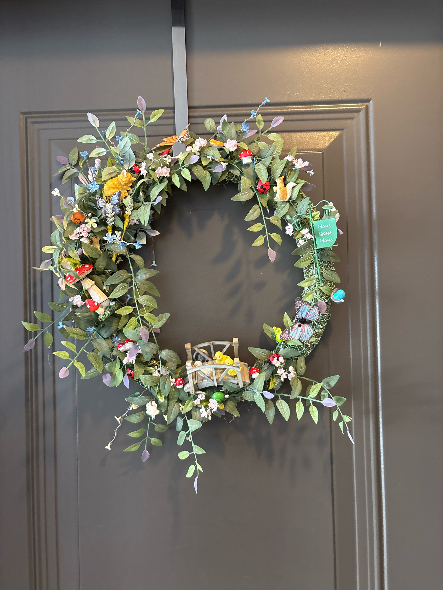 A lush, circular wreath made of artificial green leaves, tiny pink and blue flowers, and miniature decorative items hangs on a dark gray, paneled door. The wreath is adorned with several whimsical elements: red and white toadstools, ladybugs, a fox, a squirrel, a snail, butterflies, a small frog, and a miniature wooden bridge with yellow chicks. A small green sign with the words "Home Sweet Home" is attached to the wreath. The door has a subtle texture and is illuminated by overhead lighting, casting a faint shadow on the upper portion of the door. The location is the exterior of a residence in Palmetto, United States.