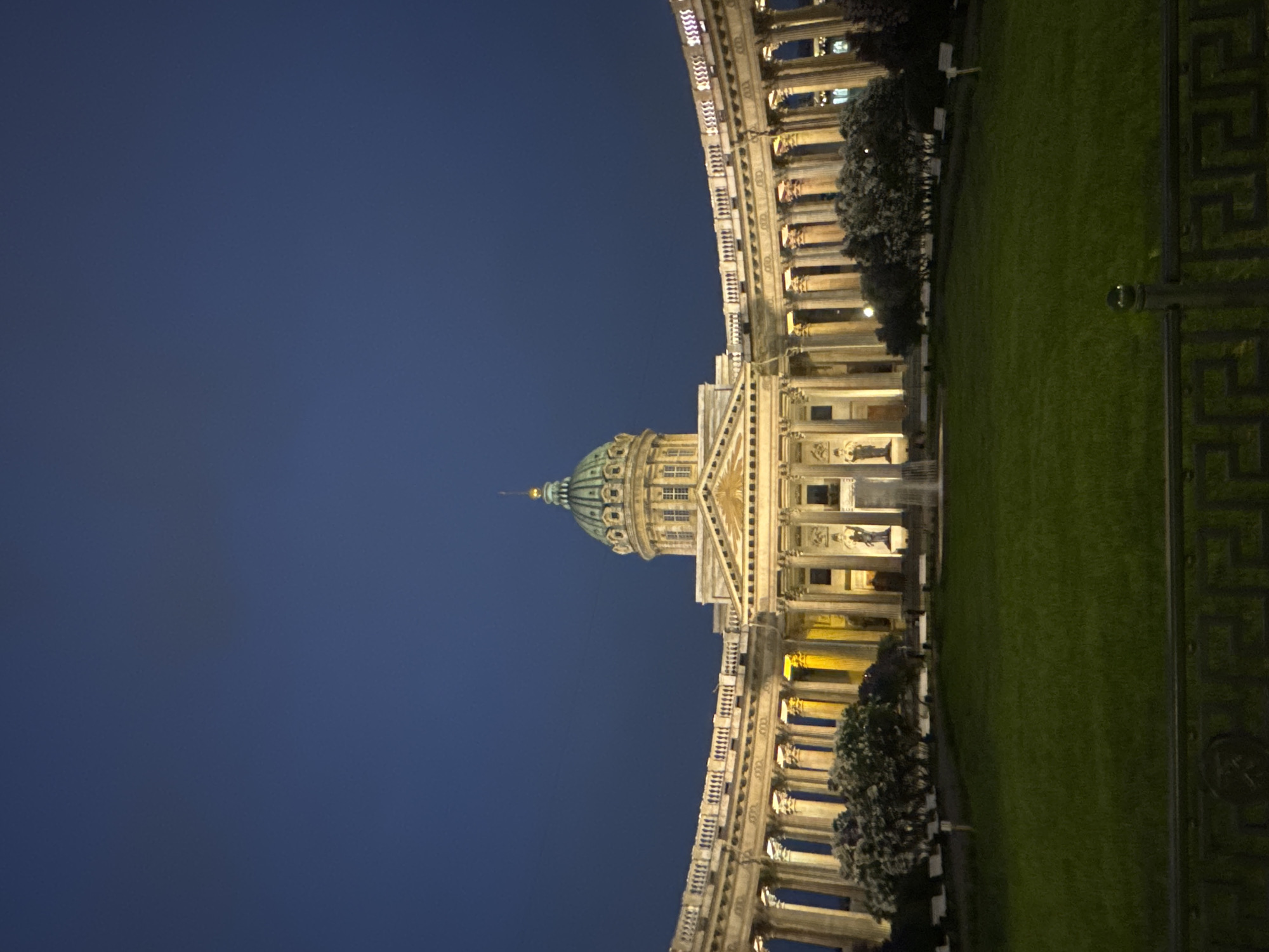 The image displays the Kazan Cathedral in Saint Petersburg, Russia, at night. The building's dome is visible along with illuminated columns and architectural details. A section of green lawn and wrought-iron fencing is on the right side of the image. The background is a dark blue sky.