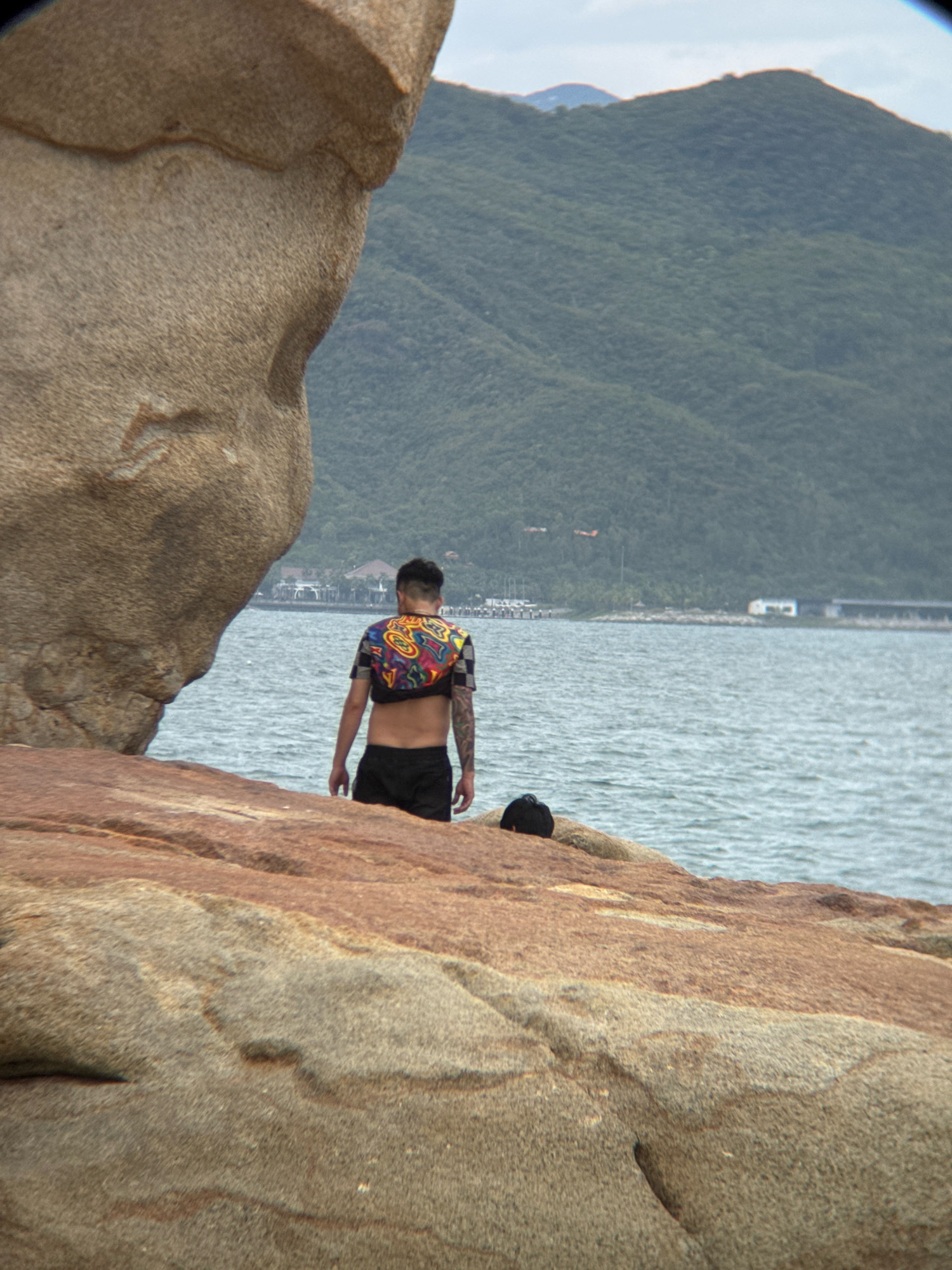 A full shot of a person, viewed from behind, stands on a rocky coastline in Nha Trang, Vietnam, facing the sea. The person, with short dark hair and tattoos on their arm, wears a cropped, colorful t-shirt and black shorts. The sea, a calm expanse of blue, meets a verdant, tree-covered mountain range in the distance. The foreground is dominated by large, weathered rocks in earthy tones. The sky is overcast.