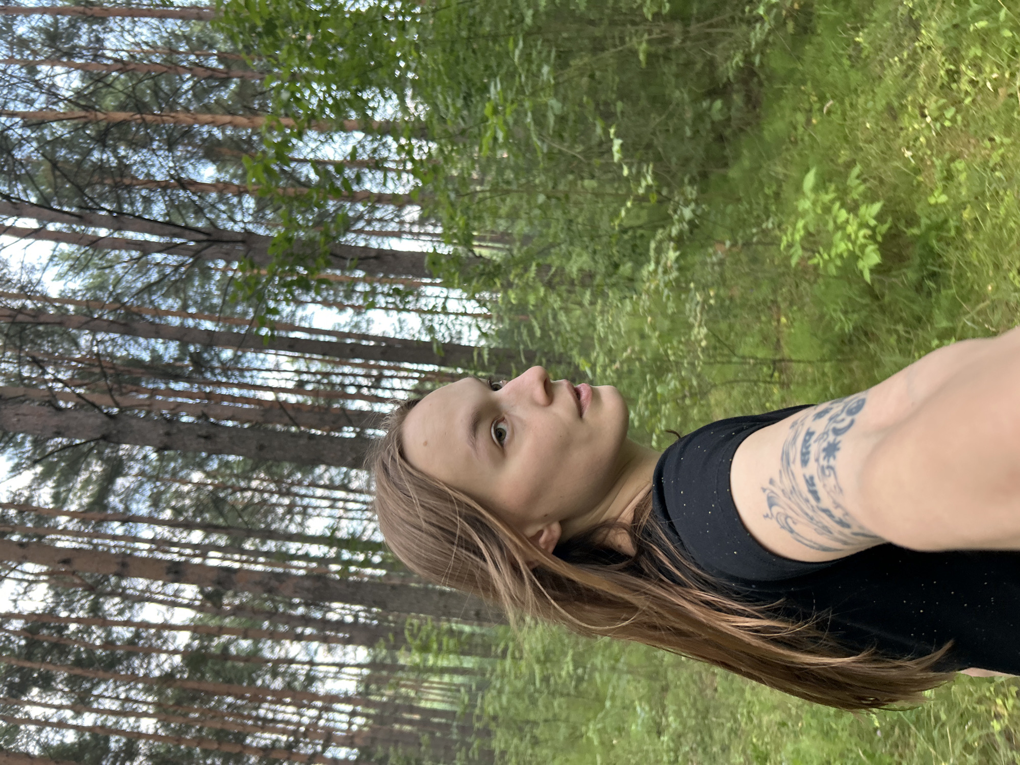 A young Caucasian woman with long brown hair and tattoos on her right arm is walking in a forest. She is wearing a black t-shirt. The background consists of tall pine trees and green foliage. The lighting suggests it is daytime. The scene appears to be a natural environment, possibly a park or woodland area.
