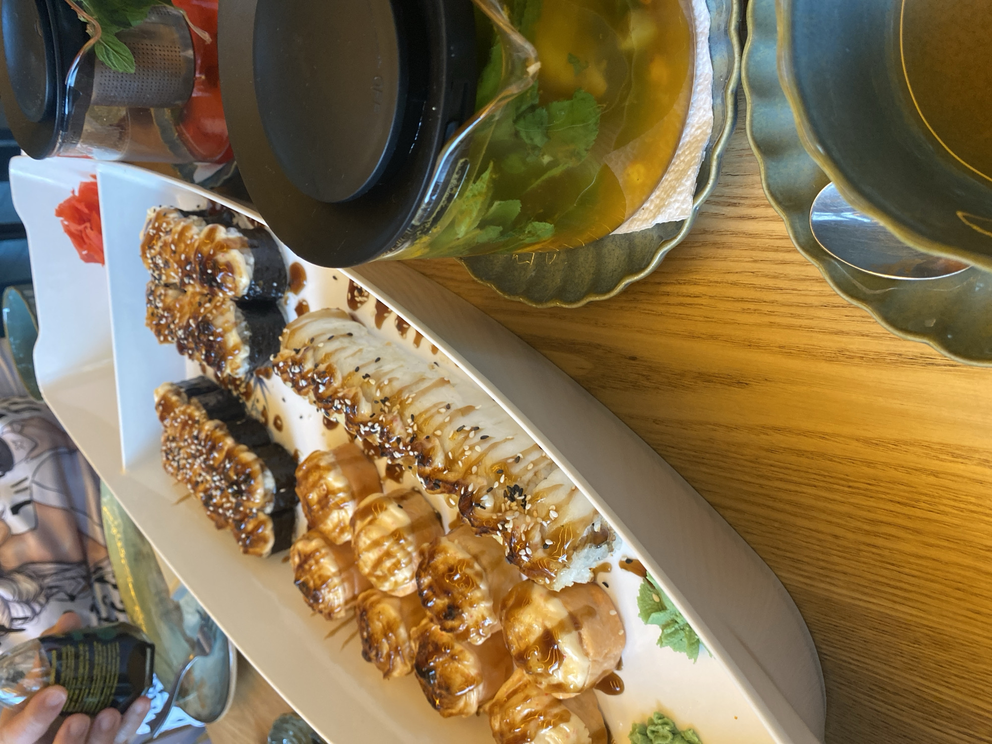 In the city of Tuymazy, Russia, a table is set for a meal. A white platter holds a variety of sushi rolls, some topped with a creamy sauce and sesame seeds, others with a glossy sauce and sesame seeds. Beside the sushi, a small pile of bright pink pickled ginger sits on the platter. To the right of the platter, a glass teapot filled with a golden liquid and green herbs steeps on a decorative plate. A black lid is on the teapot. A person's hands are visible in the upper left corner, holding a small glass bottle. The table surface is made of wooden planks. In the foreground, two dark teal bowls with scalloped edges are visible, one with a spoon inside.