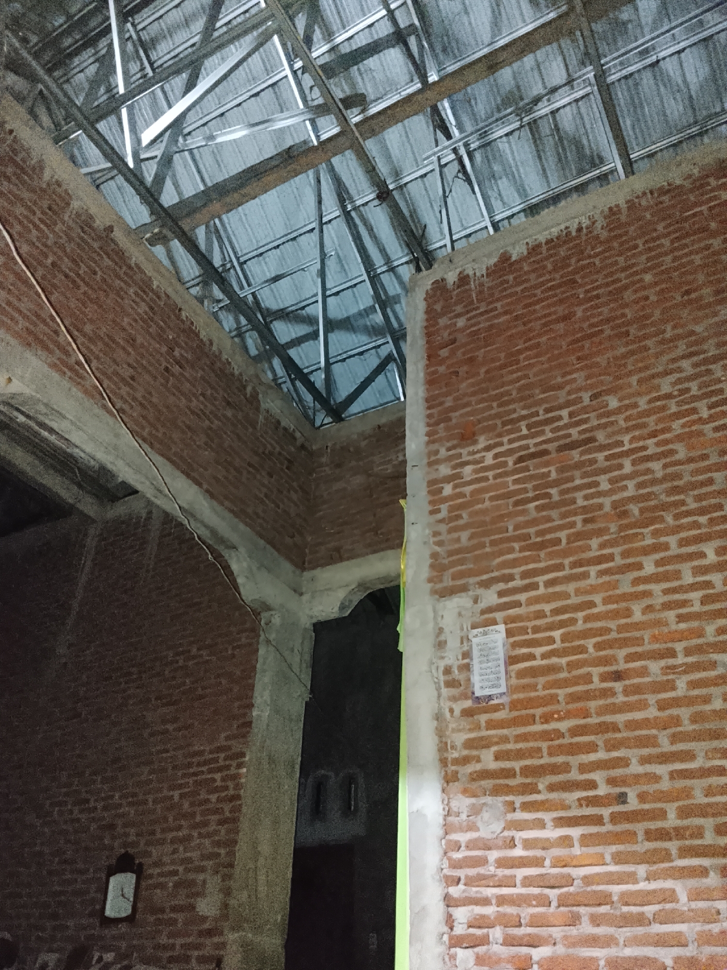The image shows the interior of a building under construction, with unfinished brick walls and a metal roof frame. A clock is visible on the left wall. The lighting is dim.
