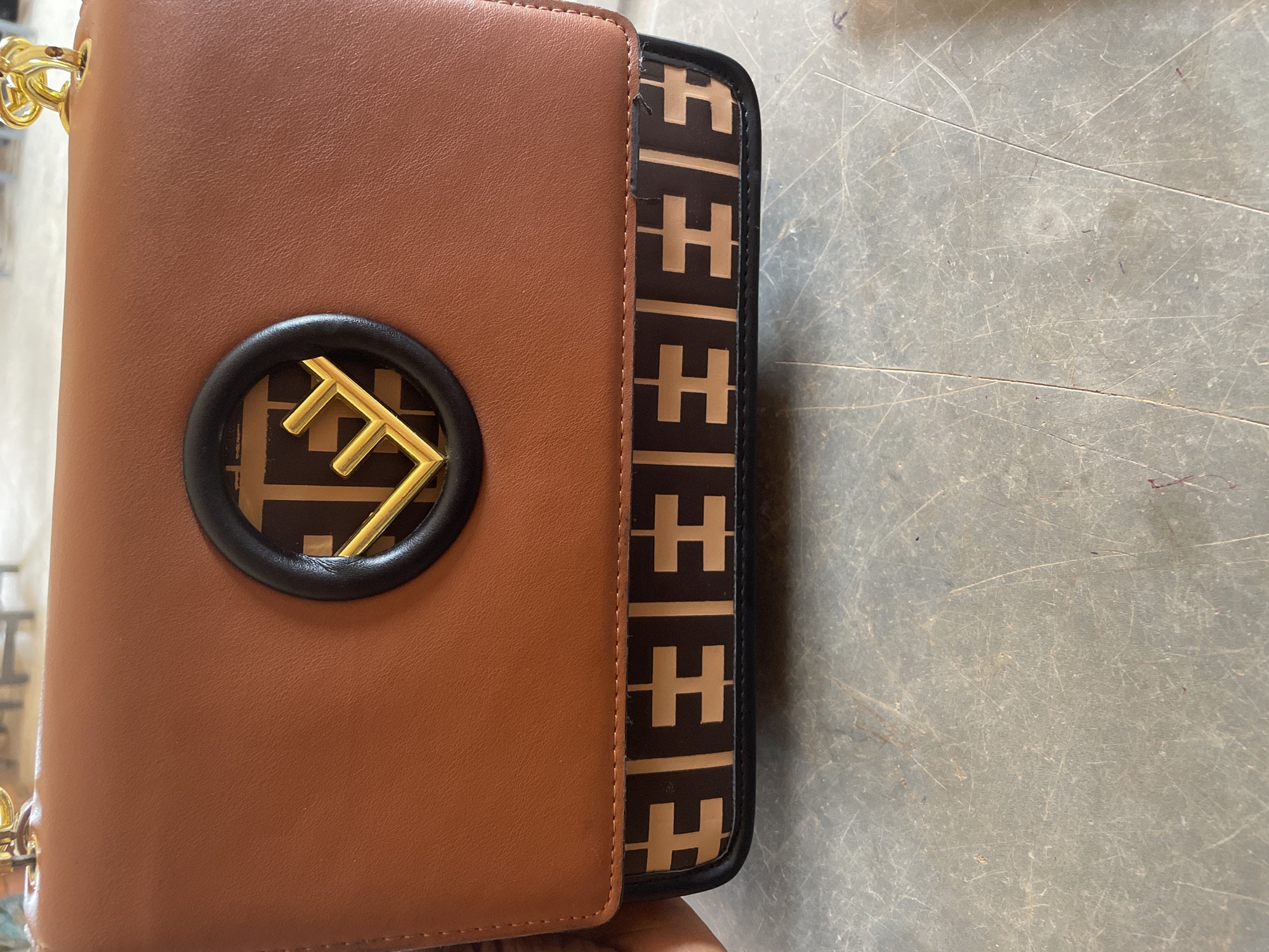 This media file showcases a close-up view of a brown handbag with a distinctive "FF" logo in gold, set against a patterned strip of the same logo in brown and beige. The bag appears to be placed on a textured, grey surface with visible scratches and markings, suggesting an indoor setting, possibly a workshop or a storage area. The lighting is neutral, indicating it could be daytime or artificial lighting, with no strong shadows. The image focuses on the details of the handbag, highlighting its material texture and the logo's metallic sheen. There is no visible text in the image. The context suggests this item might be related to fashion or retail, possibly being presented or stored in Nigeria.