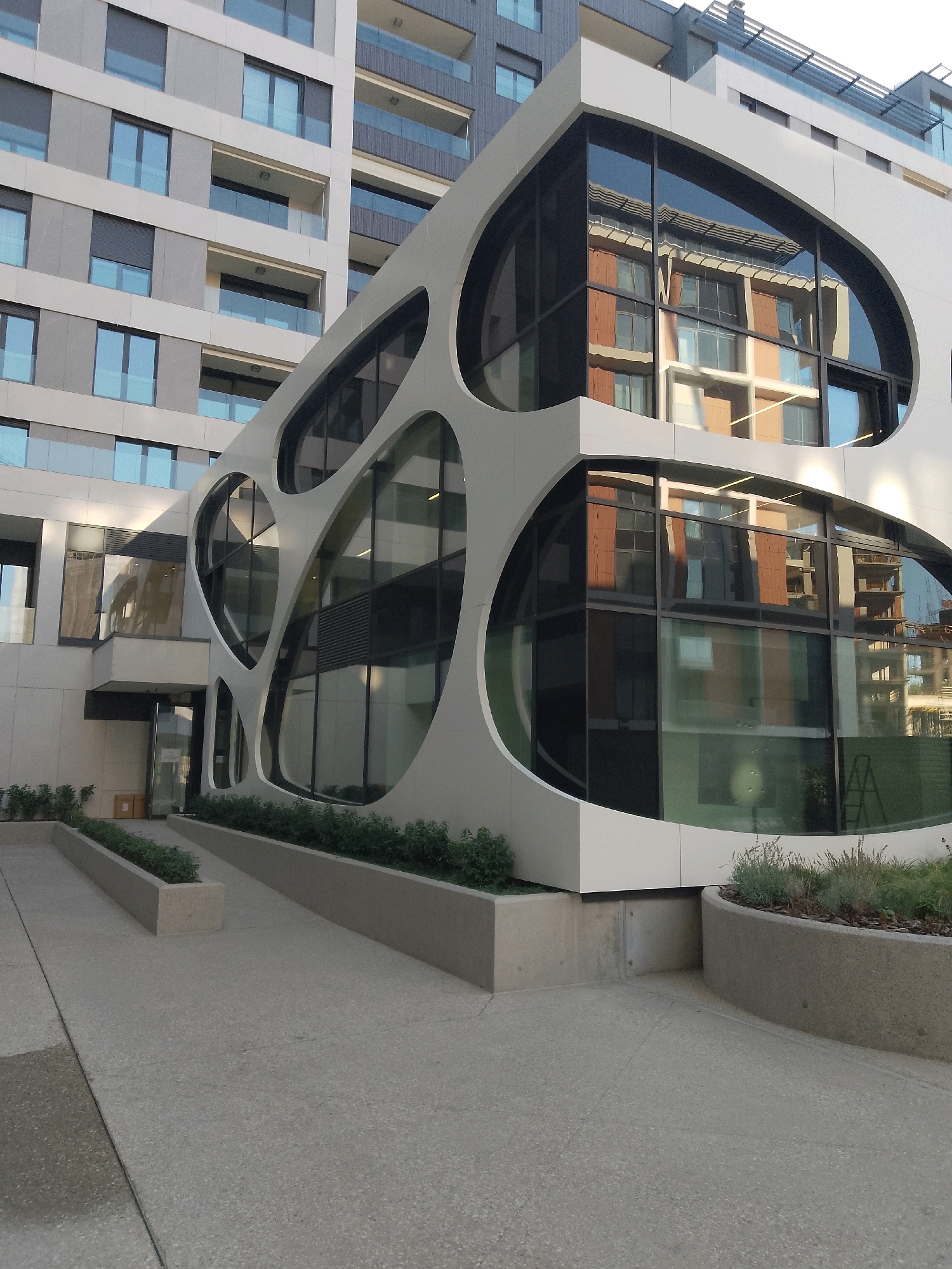 A modern building with a white, curved facade featuring irregularly shaped windows is visible in the foreground, reflecting an adjacent brick building. To the left, a taller, grey residential building with numerous windows and balconies is partially visible. The ground is paved with light-colored concrete. Several planters with greenery line the walkway and the base of the modern building. The scene appears to be an exterior view in a city.