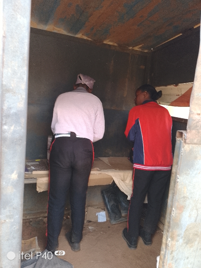 Two people stand inside a small, dilapidated structure. An older person in a light-colored sweater and dark pants with a red stripe faces a makeshift counter. A younger person wearing a red and blue jacket and similar striped pants looks towards them. The interior walls are dark and weathered, with a rusty ceiling. Debris is scattered on the ground. A pair of black boots are visible under the counter. The overall impression is one of poverty and neglect.