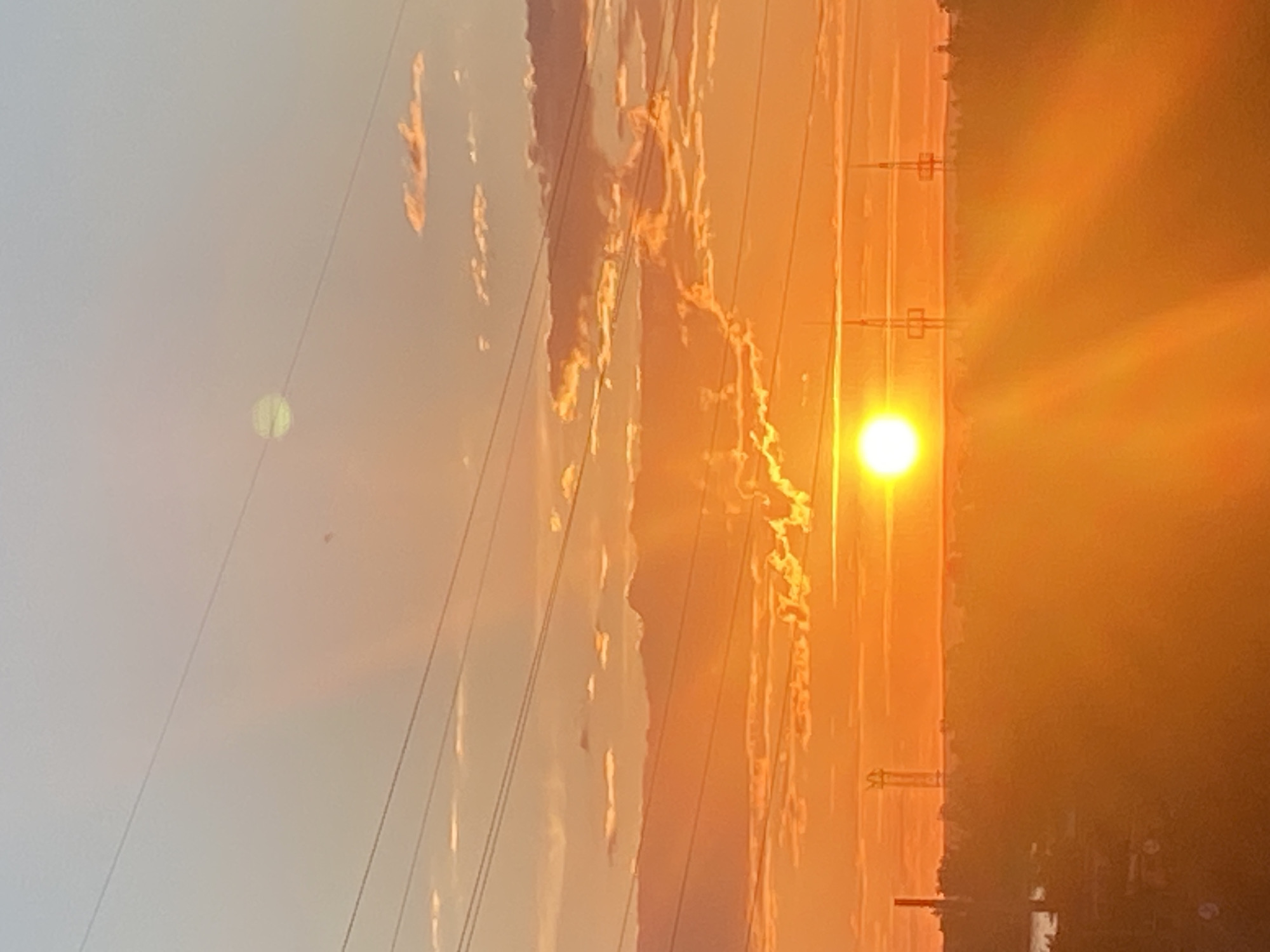 The image depicts a sunset over Novokuybyshevsk, Russia, characterized by a vibrant orange and yellow sky. Several power lines crisscross the frame from left to right. In the distance, a hazy silhouette of the city's skyline is visible, punctuated by tall industrial structures, including what appear to be towers or communication masts. The sun, a bright white orb, is low on the horizon, casting a strong glare and creating lens flares that streak across the lower portion of the image. Some clouds are scattered across the sky, catching the warm hues of the setting sun. The overall atmosphere is one of an industrial urban landscape bathed in the dramatic light of sunset.