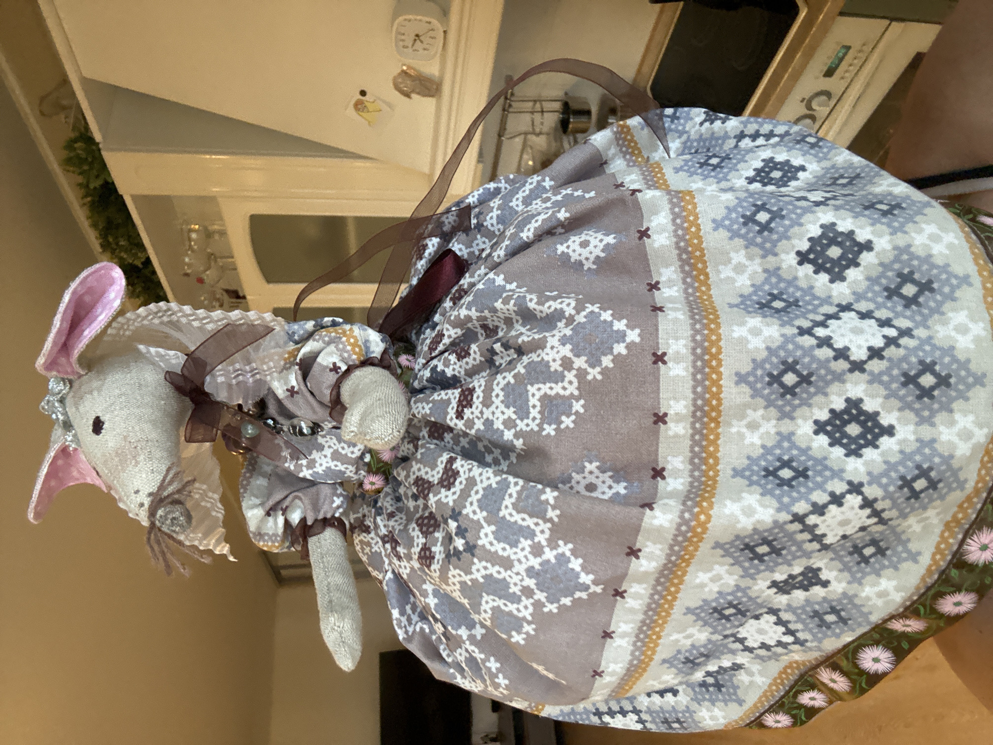 A cloth doll mouse with pink ears and a grey body, wearing a patterned dress with brown ribbon accents, sits on a counter in an indoor setting. The dress has a complex geometric pattern in shades of grey, white, and brown, with a floral trim at the bottom. The background includes a white kitchen cabinet and countertop, a clock, and a glimpse of an oven. The city and state are Unknown, Unknown.