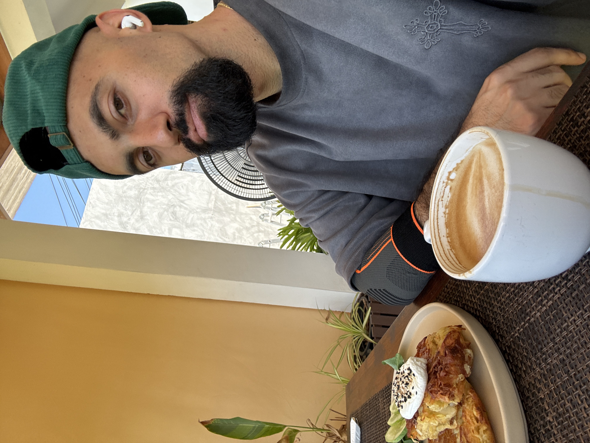 A man with a shaved head, a beard, and a green baseball cap is sitting at a table outdoors. He is wearing a gray sweatshirt with a cross design on the sleeve and a black and orange arm sleeve. In front of him is a plate of food, possibly hash browns, topped with a dollop of white cream and seeds, and a wedge of lime. To the right of the plate is a white mug filled with coffee and foam. The background shows a light orange wall, some green plants, and a white pillar. The man appears to be in Ko Samui District, Thailand, based on the implied tropical environment.