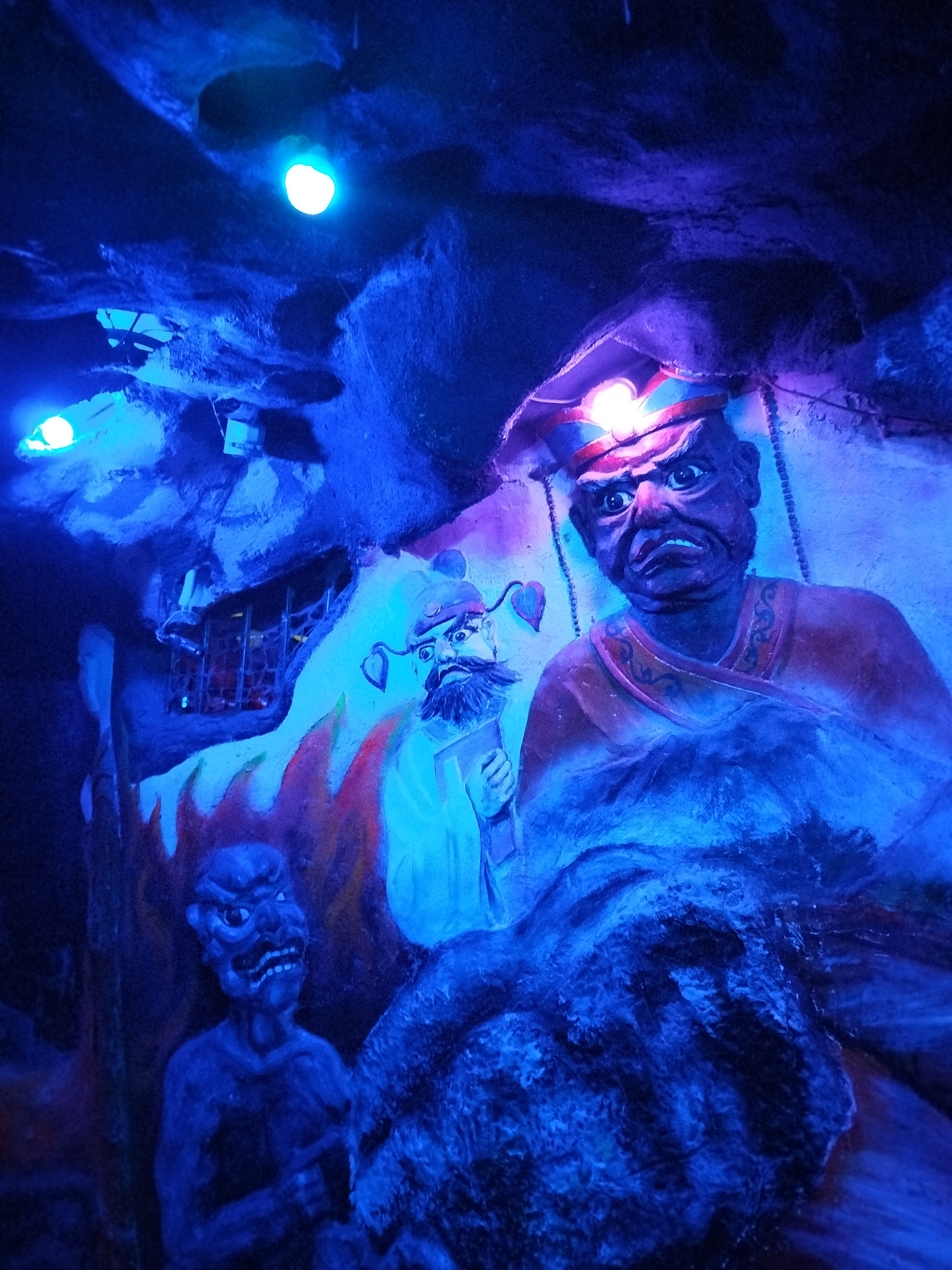 The image shows a dimly lit, cave-like interior with sculptures of demonic figures. The dominant figure is a large, stern-faced statue wearing a hat with a light on top, and an orange robe. To the left is a smaller demon figure with bare chest and an angry expression. Behind the figures, there is a painting of another bearded figure, flames, and a window. The ceiling is uneven with a few lights. The location is Unknown, Unknown.