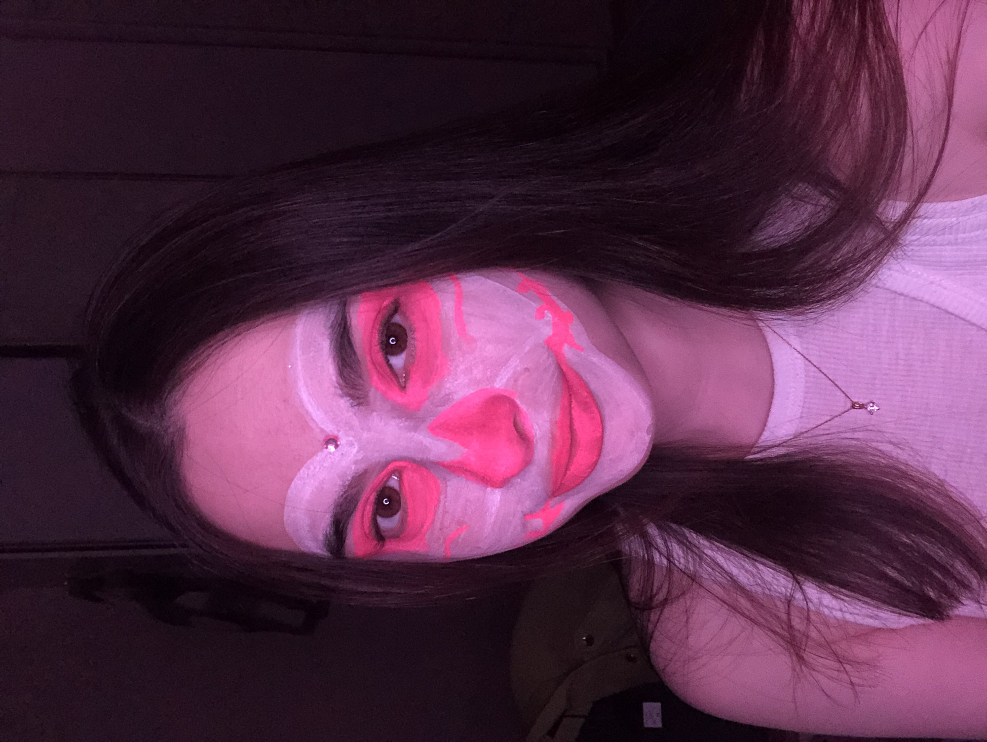 A young individual with long, dark hair is depicted in a close-up, sideways portrait, their head oriented to the left. Their face is covered in elaborate artistic makeup, featuring a white base with vibrant neon pink patterns accentuating the eyes, nose, and lips, creating a stylized, abstract design. A small, shiny gem is visible near their right eyebrow (viewer's left).
The subject looks directly at the camera with a calm expression. They are wearing a white ribbed tank top, and a thin necklace with a small, sparkling pendant is visible around their neck. The scene is illuminated by strong artificial pink and purple lighting, which casts a distinctive hue over the subject and emphasizes the neon colors of the face paint. The background is dark and indistinct, suggesting an indoor setting. The image captures the individual posing to showcase the dramatic makeup. No text is visible.