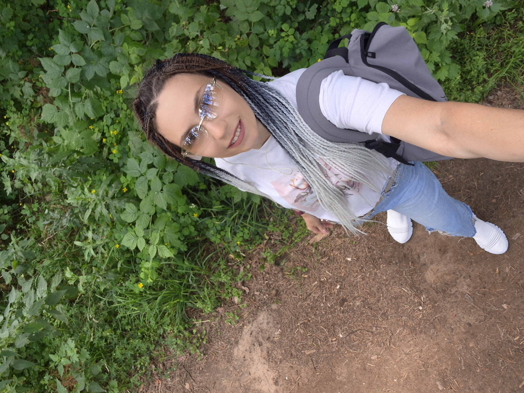 A high-angle, full-body selfie captures a woman standing on a dirt path in a wooded area. She is wearing a white t-shirt with a graphic print, ripped light-wash jeans, and white sneakers. She has long, braided hair with ombre coloring from dark brown to gray and white. She is also wearing rimless, tinted sunglasses. A gray backpack is slung over her right shoulder. The background is filled with lush green foliage, suggesting a natural or park-like environment, possibly in the city of Chaikovsky, Russia, given its reputation for having many green spaces and recreational areas.