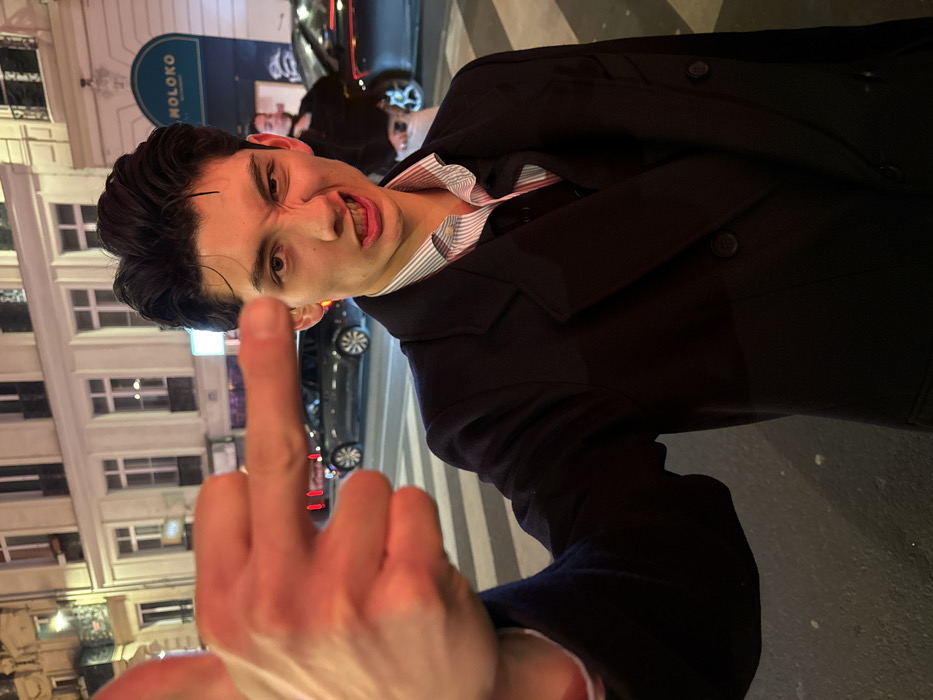 A man with dark hair and a dark suit, showing his middle finger with a grimace on his face, stands outdoors. He's wearing a striped shirt. A building with windows is in the background. A car is parked nearby. A sign with the word "MOLOKO" is visible on the building. The image is taken at night.