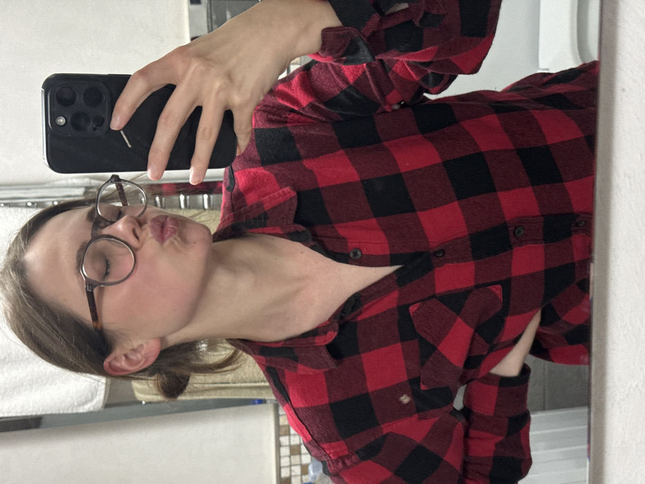 The scene depicts a mirror selfie taken by a female subject in a bathroom located in Cava de' Tirreni, Italy. The subject is shown from the mid-torso up, wearing a red and black buffalo plaid long-sleeved flannel shirt with the top button undone. Her hair is dark brown and pulled back in a bun. She wears round, dark-rimmed eyeglasses. Her eyes are closed, and her lips are pursed in a "kissing" gesture. Her right hand, holding a dark-cased smartphone (consistent with an iPhone featuring a three-lens camera system), is visible in the upper right portion of the frame, angled towards the mirror to capture the reflection. The subject's left hand is placed on her hip. The background behind the subject's reflection reveals a wall with a metal towel rack holding a white towel on the left, and a tiled wall featuring small, light-colored square tiles on the right. White bathroom fixtures are partially visible at the bottom edge of the frame.