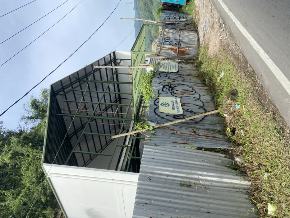 The image shows a partially constructed building in Soreang, Indonesia.  The building is made of metal framing and is covered with a white material. The building is located on a hillside, and a corrugated metal fence surrounds it. There is a sign on the fence that says "utamakan factor keselamatan" which translates to "prioritize safety factors." There is a dirt road next to the fence and a paved road to the right. Green vegetation covers the hillside in the background.  The coordinates (latitude: -7.037, longitude: 107.497) correspond to a location in Soreang, Indonesia.
