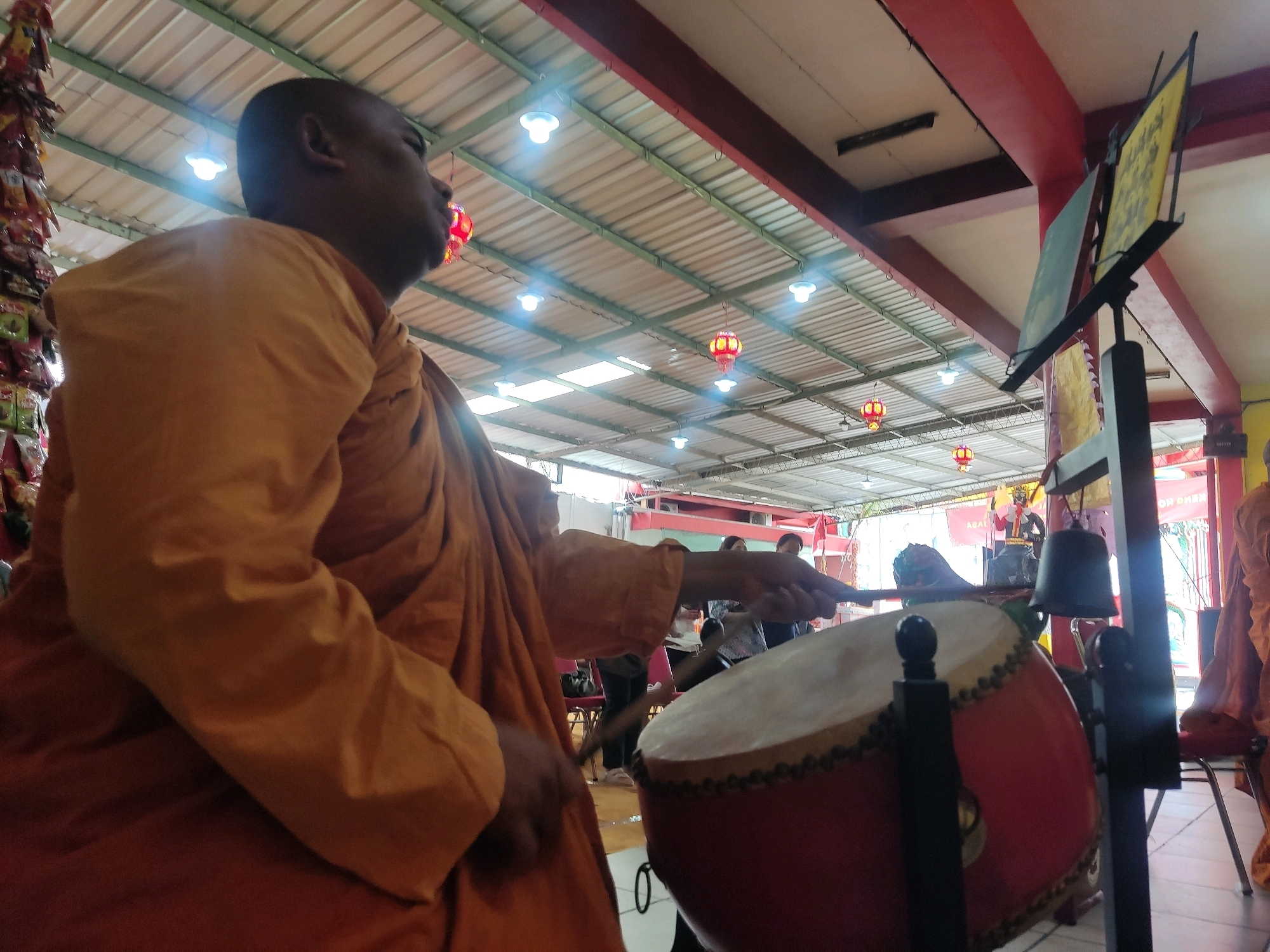 A man wearing an orange robe is playing a large red drum with a drumstick. He is positioned in the foreground, looking towards the right of the frame. Behind him, there is a drum stand with a bell hanging from it. In the background, there are several people, chairs, and what appear to be religious decorations, including lanterns. The scene is set indoors, under a corrugated metal roof supported by green metal beams. Red structural beams are visible on the ceiling. The overall lighting suggests daytime. The image was taken in Magelang, Indonesia, at a cultural or religious event.