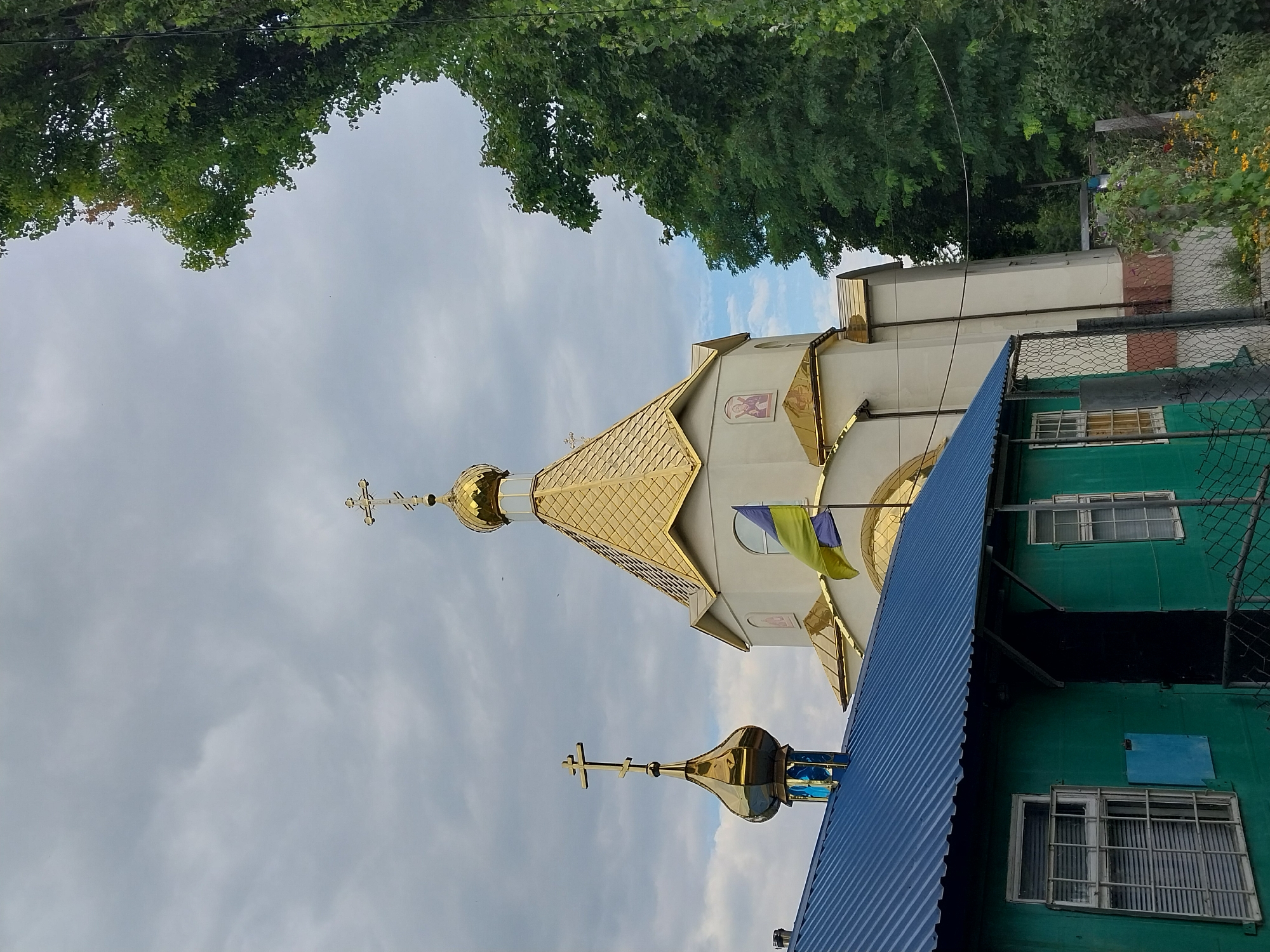 The image captures a scene in the city of Doslidne, Ukraine, dominated by a white church with golden domes. In the foreground, a small, green building with a blue corrugated metal roof is visible. The building has at least two windows, one of which is secured with metal bars. A chain-link fence separates the green building from the church. A Ukrainian flag, horizontally striped in yellow and blue, flies on a pole in front of the church. Lush green trees line the right side of the image, with a cloudy sky overhead. The overall impression is one of a religious structure and its surroundings under an overcast sky.