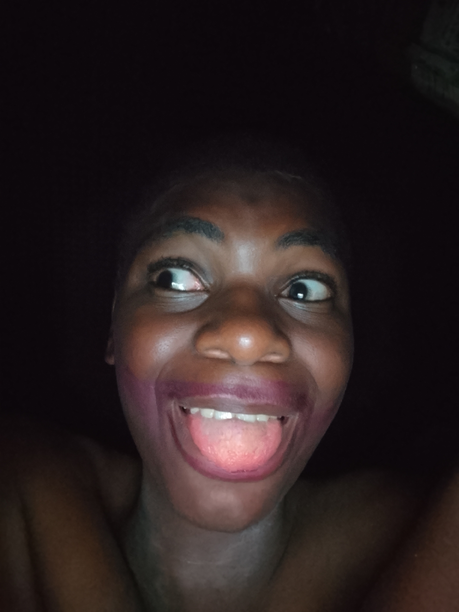 This close-up portrait, set in Nkpor, Nigeria, features a dark-skinned individual making a playful or silly facial expression against a completely dark background, suggesting a low-light indoor setting or nighttime. The subject's face fills most of the frame, with their shoulders partially visible at the bottom.
The person has wide, expressive eyes, with the left eye (viewer's right) looking towards the upper left corner and the right eye (viewer's left) looking towards the upper right corner, creating a cross-eyed effect. Their mouth is open wide, revealing white teeth and a pink tongue sticking out. There is smudged purple or dark pink lipstick or makeup around the mouth, extending onto the skin around the lips, chin, and cheeks, contributing to the comical appearance. The overall emotion conveyed is one of humor, amusement, or silliness. No specific objects or text are visible in the image beyond the person. The lighting emphasizes the facial features against the dark surroundings.