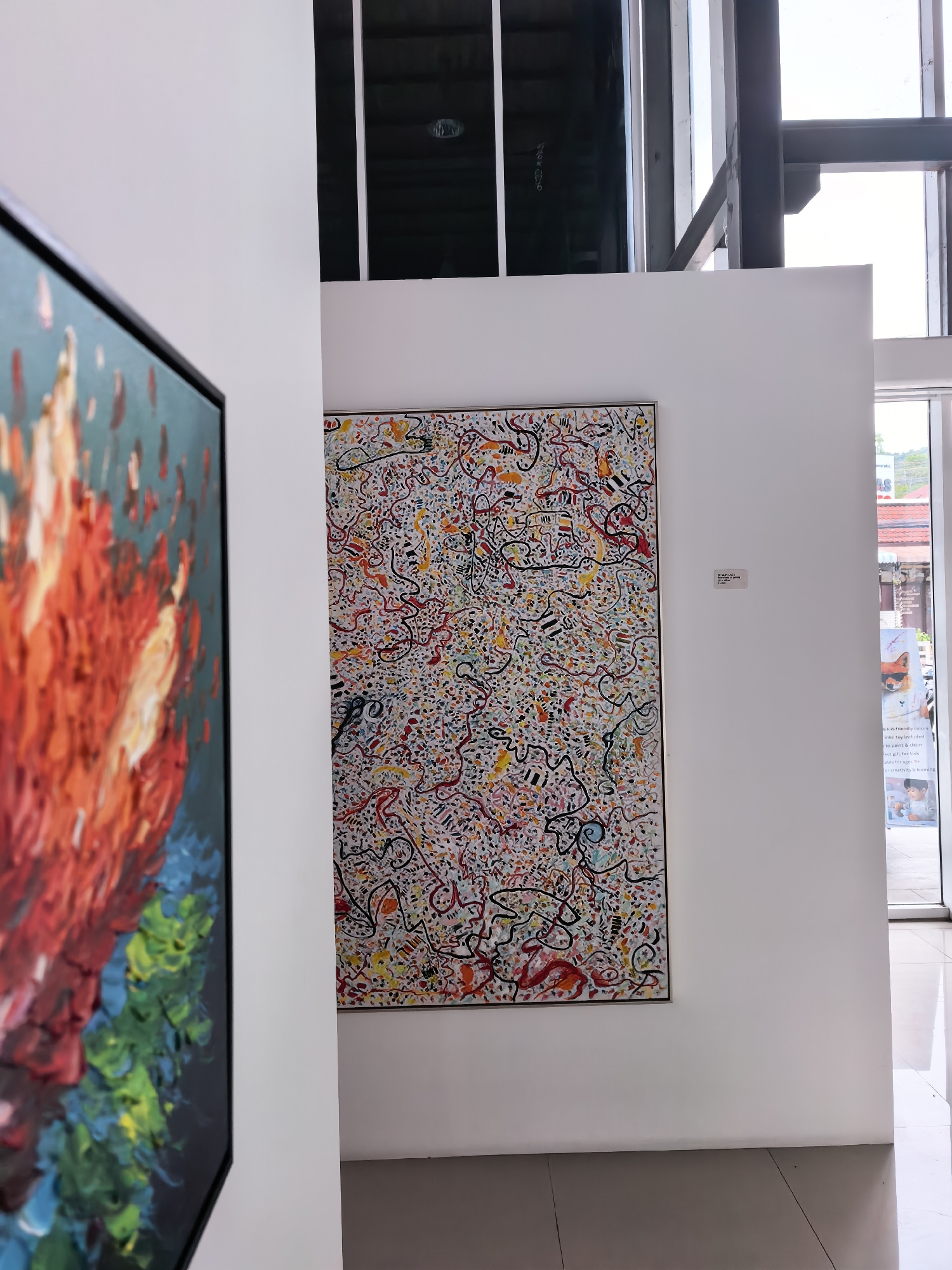The image depicts the interior of an art gallery or exhibition space. Two paintings are visible. On the left, a portion of a painting with bold strokes of red, orange, yellow, green, and blue is shown, rendered in thick impasto. To the right, a larger, framed abstract painting is displayed on a white wall. This painting is characterized by a white background covered with a dense network of thin, multicolored lines, dots, and small shapes in red, black, yellow, blue, and orange. A small label is affixed to the wall next to the abstract painting. The environment includes white walls, a dark ceiling with visible structural elements, large windows or glass doors, and a tiled floor. Outside the window, hints of an outdoor environment with greenery and buildings are visible, suggesting an urban or semi-urban setting. The lighting appears to be a mix of natural light from the windows and artificial light. The location is identified as the city of Ko Samui District, Thailand. There are no people clearly visible in the image, but a poster with images of children and products can be seen through the glass.