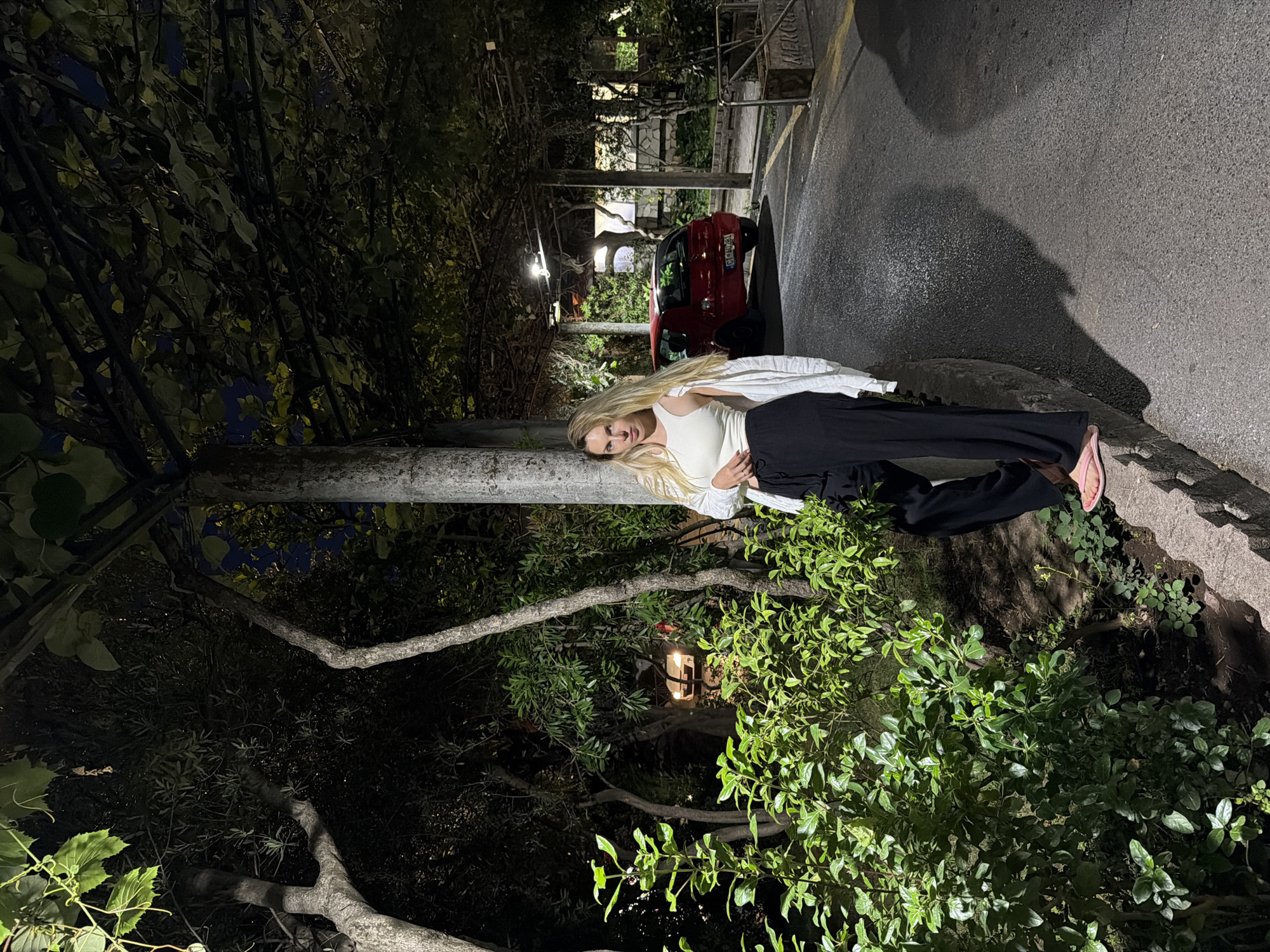 In the city of Budva, Montenegro, at night, a blonde woman with long hair stands leaning against a concrete pillar under a vine-covered pergola. She is wearing a white tank top, black wide-leg pants, and pink slides. To her left, lush green foliage borders a stone wall. To her right, an asphalt road curves away, with a small red car parked on it. The scene is dimly lit by artificial lights, casting shadows and highlighting the textures of the plants and road.