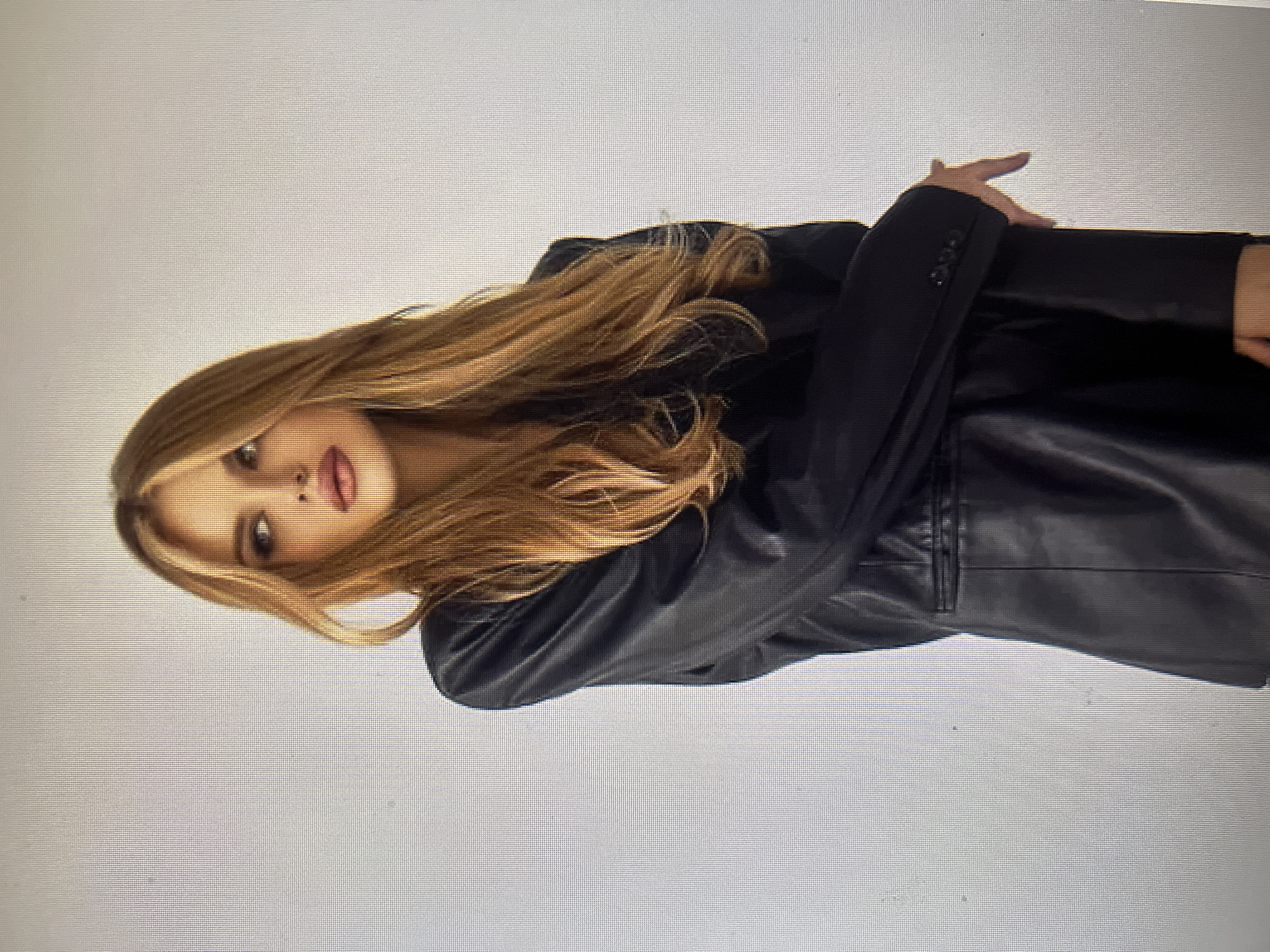 A young woman with long, wavy, blonde hair and fair skin is posing for a photograph. She is looking to her left, with her head tilted slightly. She has full lips and is wearing dark eye makeup. She is wearing a black leather blazer with a lapel and buttons on the cuffs. Her arms are crossed in front of her. The background is a plain, light gray wall. The image does not contain any information about the location in the city of Qarağandı city administration, Kazakhstan.