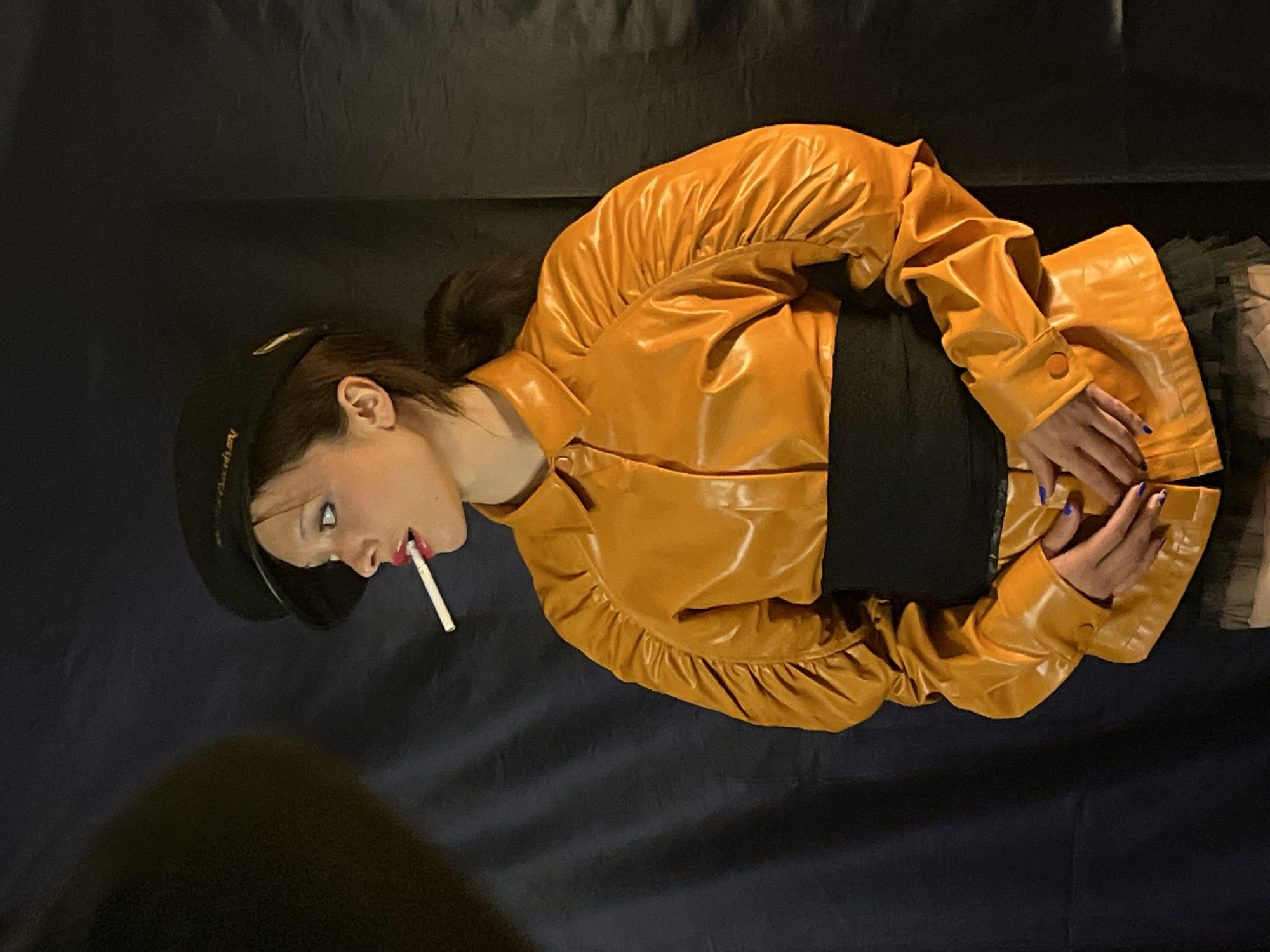 A person with dark hair pulled back into a bun, wearing a black hat and a yellow, leather-like jacket, lies on a dark blue surface. The person has a cigarette in their mouth. Their hands are crossed in front of them. The image does not show details that would specify its location.