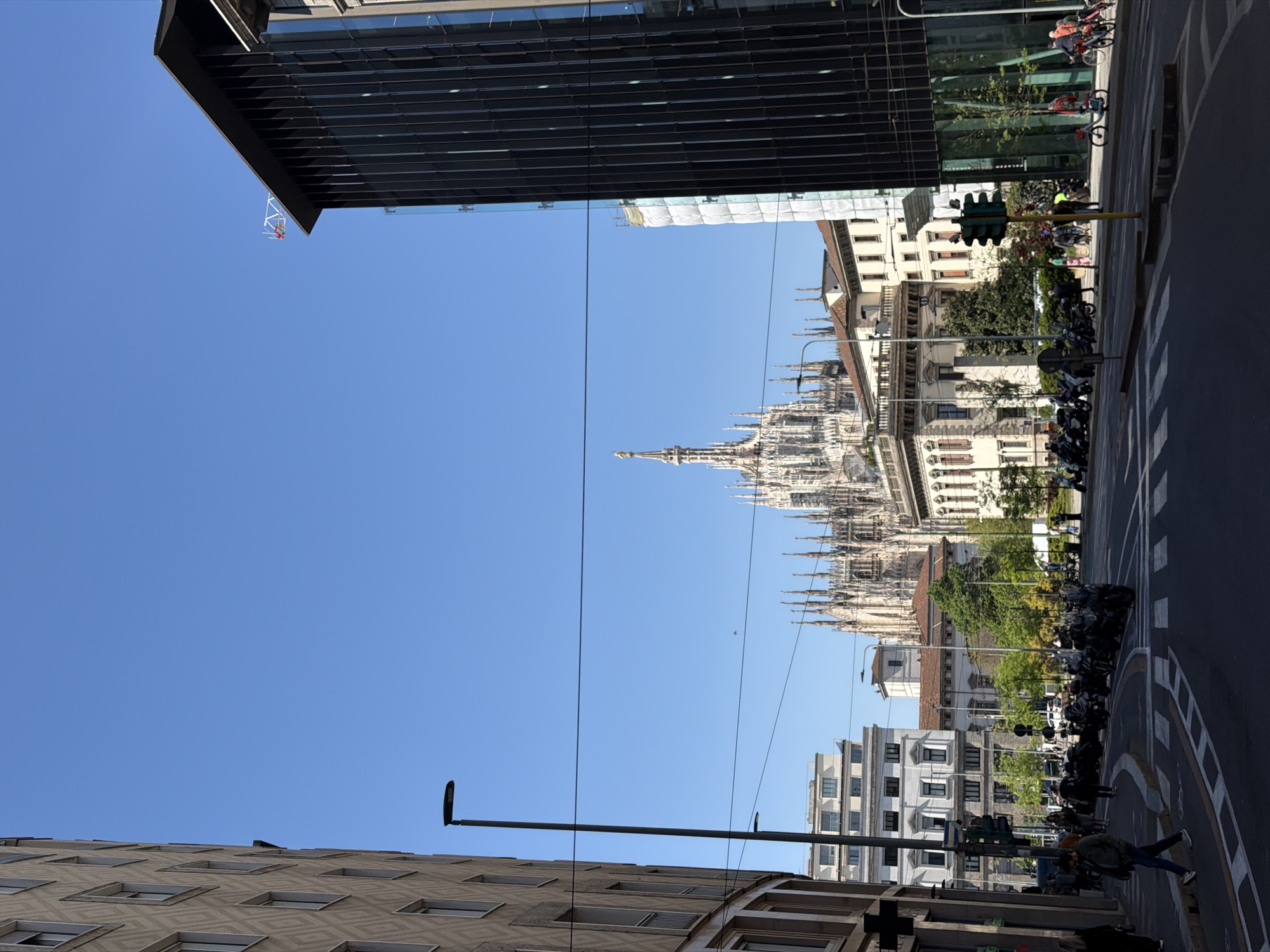 The image captures a street scene in Milan, Italy, featuring the Milan Cathedral (Duomo di Milano) in the background. The sky is clear and blue. The scene is framed by buildings on either side, with a modern, dark-colored building on the left and a lighter, patterned building at the bottom. Street-level details include parked motorcycles lining the road. A traffic light is visible, and there are pedestrians present. The road has marked crosswalks. Overhead wires are also present in the scene.