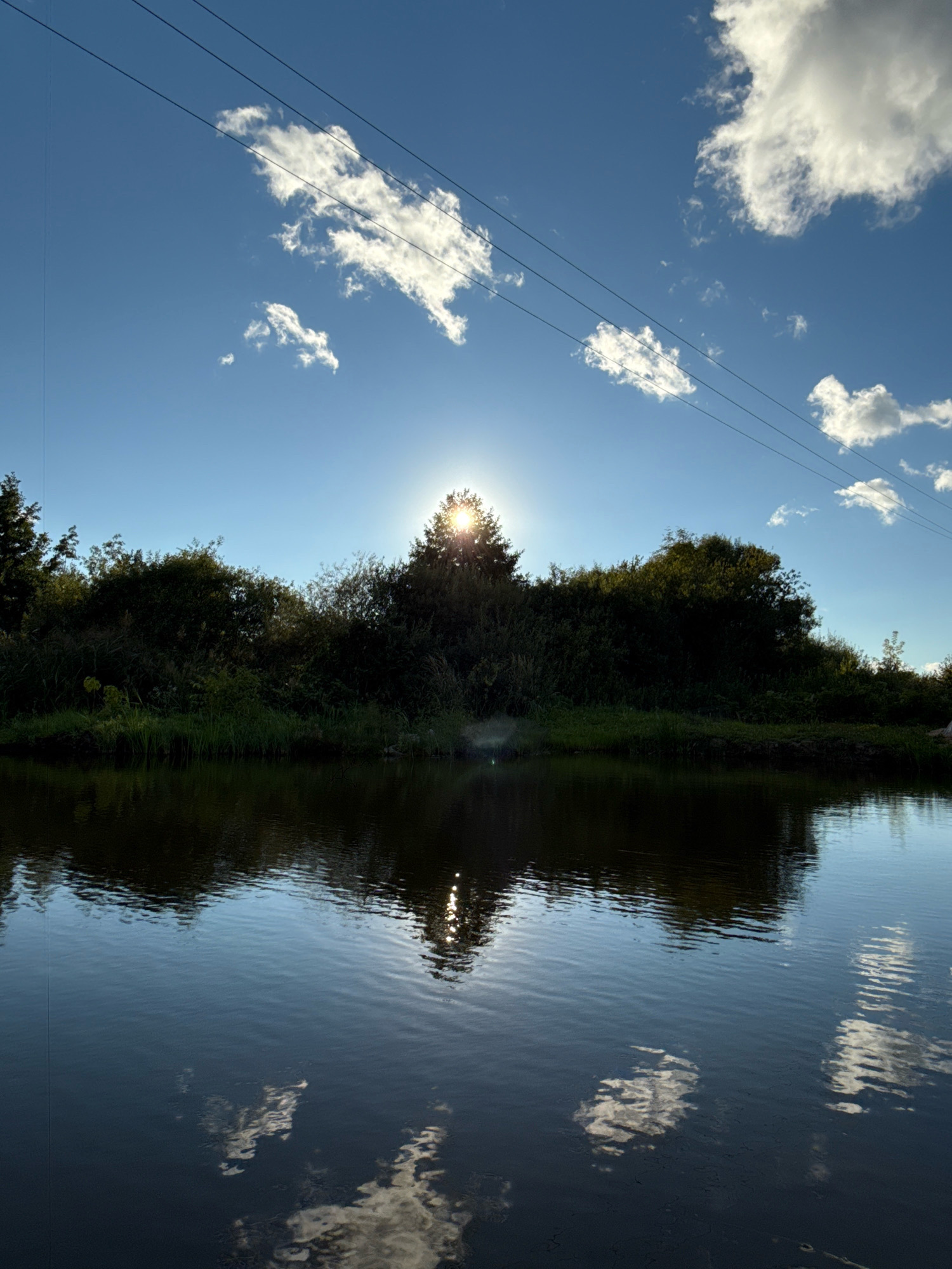The image depicts a serene natural scene with a body of water in the foreground and a dense line of trees and foliage forming the background. The sun is partially obscured by a tree on the right side, casting a bright glow and creating a sunburst effect. The sky above is a vibrant blue with scattered white clouds. Power lines are visible stretching across the upper portion of the image. The water's surface reflects the sky, clouds, and the surrounding vegetation, creating a mirror-like effect. The overall impression is one of a peaceful, sun-drenched landscape. The location is specified as Slawharad, Belarus.