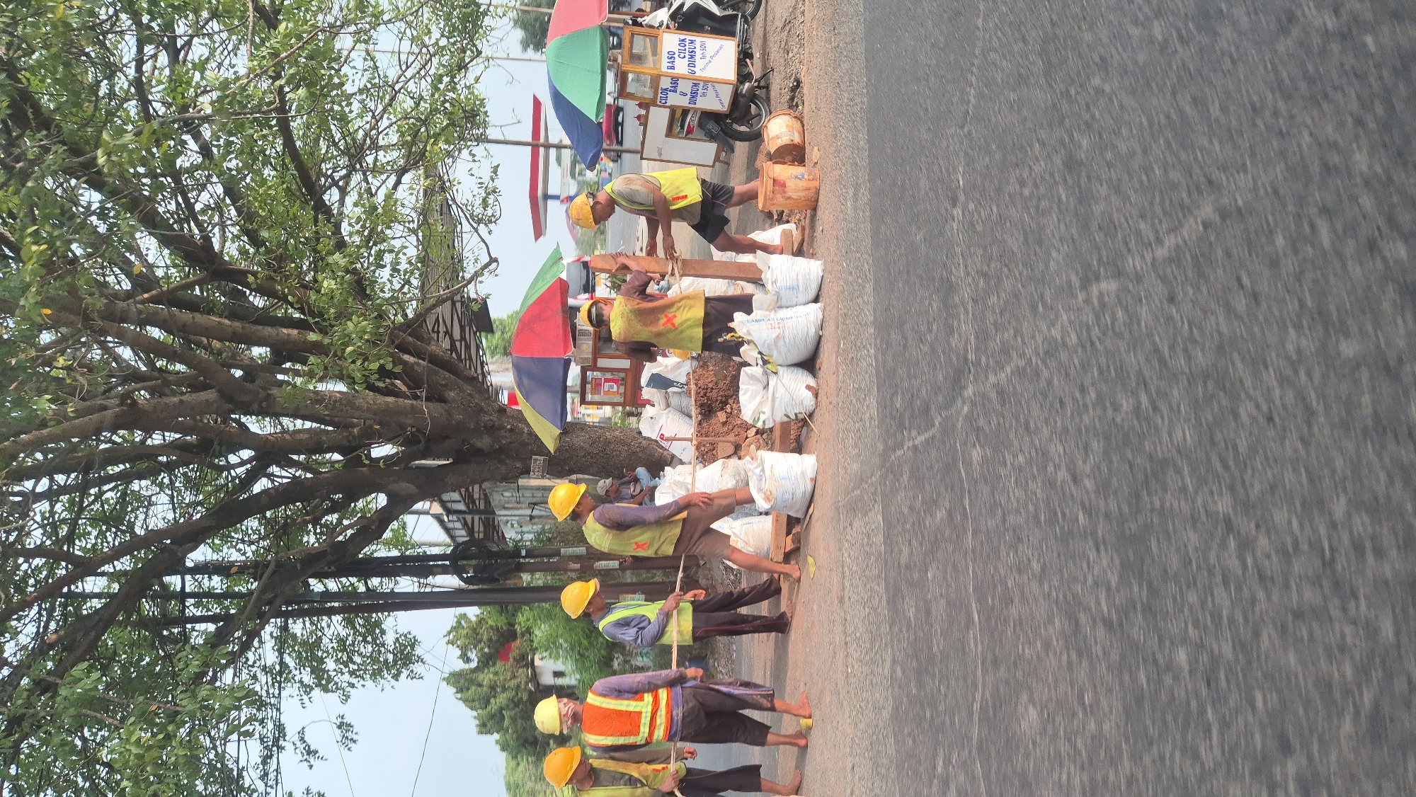 The image shows a group of construction workers on a roadside in Karawang, Indonesia, likely engaged in road repair or maintenance. Six men, all wearing yellow hard hats and reflective vests, are present. Four of them are actively working near a pile of white sacks, which appear to be filled with sand or soil, and a wooden post. One man is holding the post while another appears to be securing it. Two other workers are standing further to the left, observing. In the background, there are several colorful umbrellas, a small food stall, a motorbike, and a building structure. The scene is set under a large tree, with power lines visible behind it. The road surface is asphalt, and the lighting suggests it is daytime, possibly late afternoon due to the warm tones in the image.
