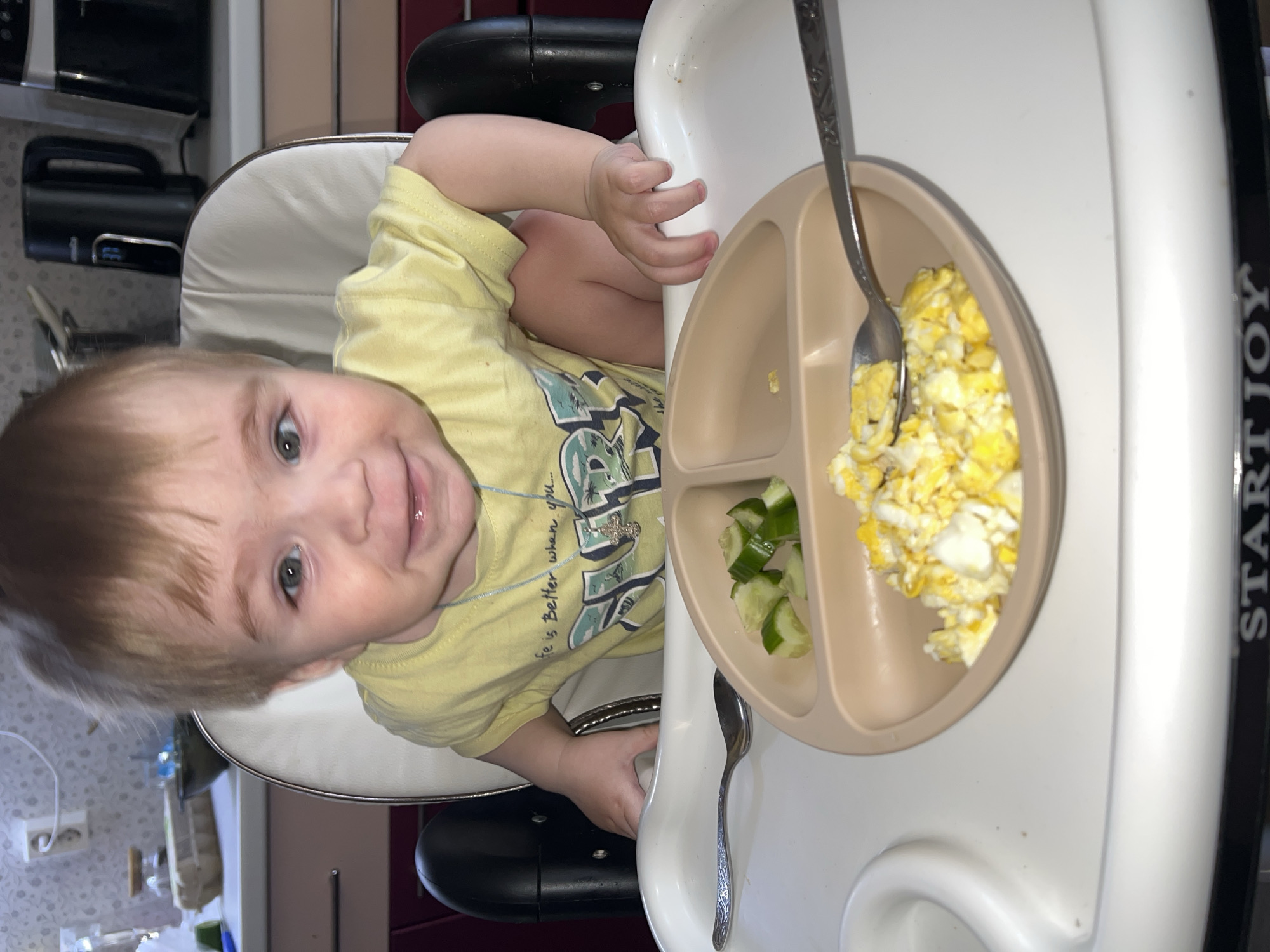A toddler sits in a high chair, angled slightly to the left in the frame. The baby wears a yellow shirt with a graphic print and a small necklace. On the high chair tray is a divided plate containing scrambled eggs and sliced cucumbers, with a spoon resting in the eggs. A second spoon lays on the tray beside the plate. The high chair has a white tray and seat, with black armrests. The brand "START JOY" is partially visible on the right side of the tray. In the background, kitchen appliances and cabinets are visible. The location is Noviy, Russia.